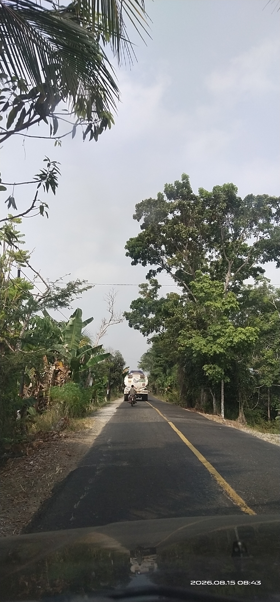 A low-angle, eye-level shot captures a road scene in Bambulung, Indonesia, under a hazy sky. The road, paved and featuring a yellow center line, curves slightly to the right and extends into the distance. On the left side of the road, dense foliage comprising various trees and plants, including banana trees, creates a natural border. On the right, a thick canopy of trees lines the road. In the middle ground, a white tanker truck with the text "PERTAMINA" visible on its side travels away from the camera, followed by a motorcycle carrying a single rider. The foreground shows the hood of a vehicle, reflecting the surrounding environment. The timestamp "2026.08.15 08:43" is present in the bottom right corner.