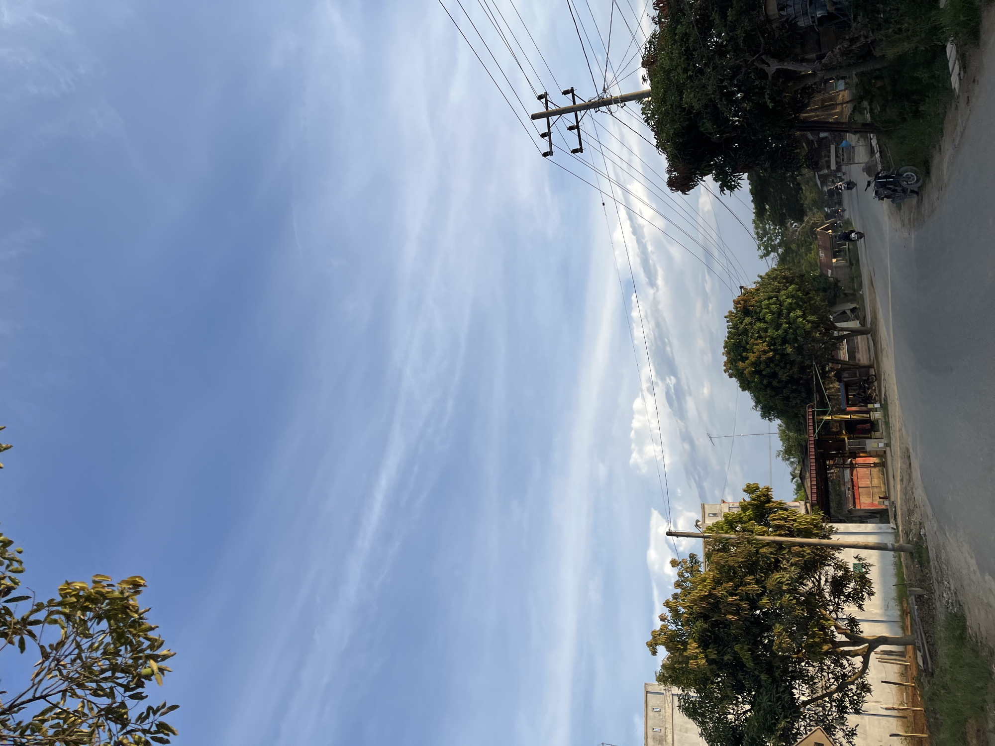 The image depicts a street scene in Durian, Indonesia, under a bright, partly cloudy sky, suggesting it is daytime. The perspective is from a low angle, looking upwards towards the sky and the elements along the road.
The primary subjects are the sky, a road, trees, and man-made structures. The sky is a vibrant blue with wispy white clouds streaking across it. A prominent feature in the sky is a utility pole with numerous wires extending from it, typical of an urban or semi-urban environment.
Along the right side of the image, a paved road runs diagonally. There are several trees lining the road, with lush green foliage. Some of these trees are quite mature with thick trunks and branches. Behind the trees, various buildings and structures are visible. Some appear to be small shops or houses with corrugated metal roofs and simple facades.
Several motorcycles are present on the road. One is parked on the side of the road, and others are seen in motion further down the street, indicating normal daily activity. No people are clearly discernible.
Notable details include the presence of electrical wires, the type of trees, and the style of buildings, which are consistent with a tropical or Southeast Asian setting. The lighting suggests it is likely late morning or early afternoon. The overall impression is one of a typical day in a residential or commercial area of Durian. No text is visible in the image.