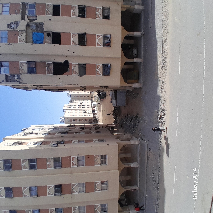 Two buildings frame a road in a vertical shot. The buildings are multi-storied with arched ground floors, and windows displaying signs of damage. Rubble lies between the buildings, and a makeshift shelter is visible. A figure stands on the roadside with a long shadow. The sky is clear and blue. The image shows signs of poverty and destruction.