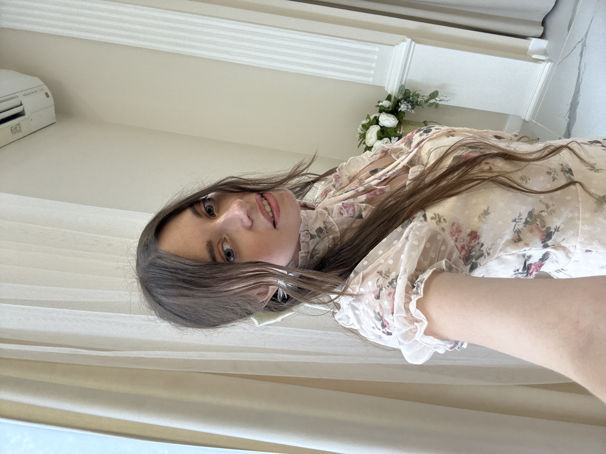 A young woman with long, brown hair and blue eyes is captured in a selfie. She is wearing a light-colored dress with a floral pattern and ruffled sleeves. The background features cream-colored curtains and a decorative white column, suggesting an indoor setting, possibly a dressing room or studio. The woman is looking at the camera with a slight smile.