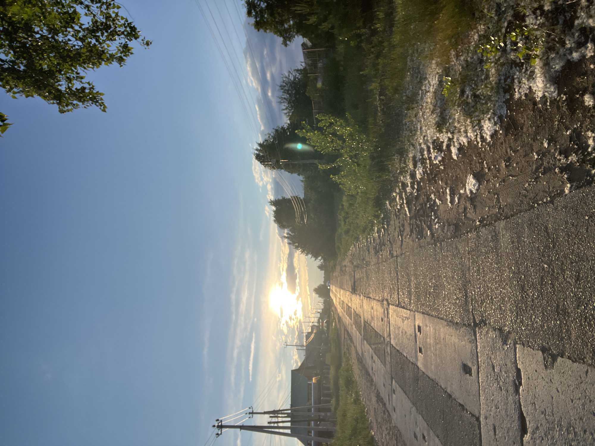 The image depicts a road stretching into the distance, with the setting sun creating a bright glare on the road surface. The road appears to be made of concrete slabs. Vegetation and trees line the sides of the road. Utility poles and power lines are visible on the left side of the image, leading towards the horizon. The sky is blue with scattered clouds. Based on the available information, the location of the image is in the city of Chaikovsky, Russia. No people are discernible in the image.