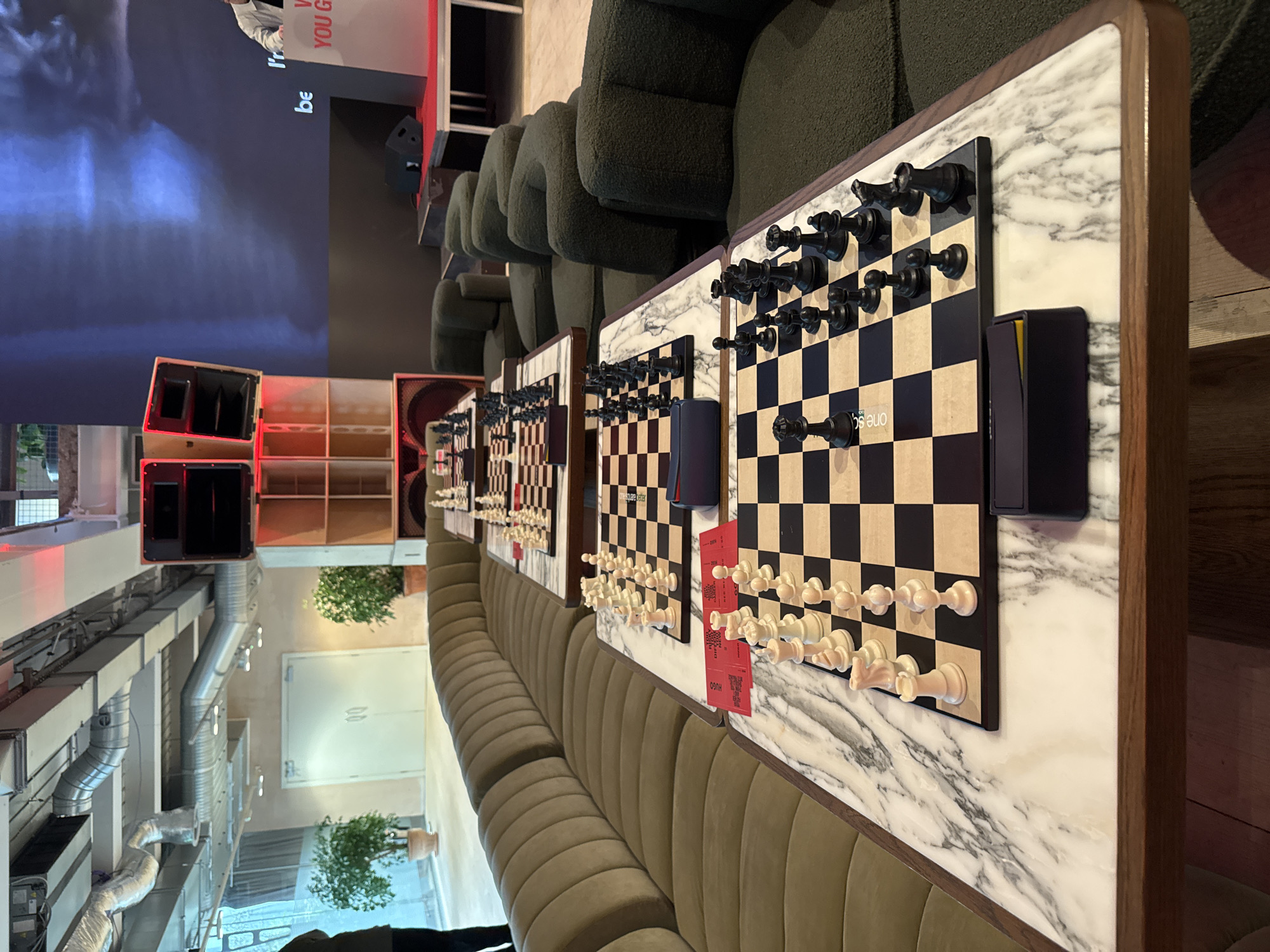 The image shows a series of chessboards set up on tables along a curved, cushioned bench in an indoor setting. The chessboards have black and white pieces arranged in various stages of play. Each chessboard has a small, rectangular object (possibly a device or marker) next to it. The tables have a marble-like surface.
Behind the tables and bench are dark green upholstered chairs. In the background, there's a shelving unit with storage spaces and red accents. To the left, there are exposed ventilation ducts and a large screen with a video playing.
The scene appears to be indoors and possibly a modern, public space or event location.
Without additional information, providing a location in London, United Kingdom is not possible.