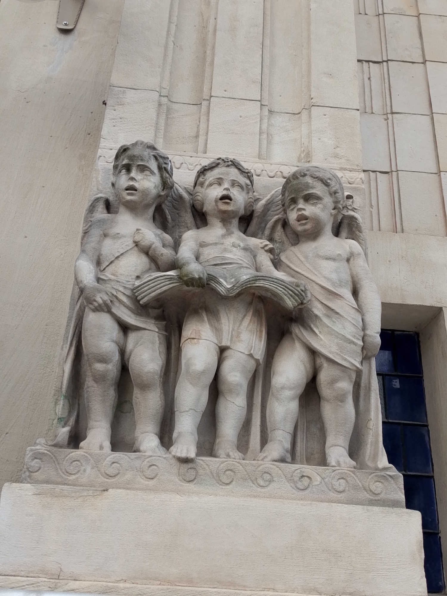 Three stone sculptures of cherubic figures, likely angels or putti, stand in a row. The figures are depicted with wings and draped clothing. The central figure holds an open book, with the figures on either side appearing to read or sing from it. The sculptures are mounted on a stone pedestal with decorative scrollwork. The background consists of a textured stone wall and a window with dark blue panes. This sculpture is located on the facade of the Ossolineum National Institute in Wrocław, Poland.