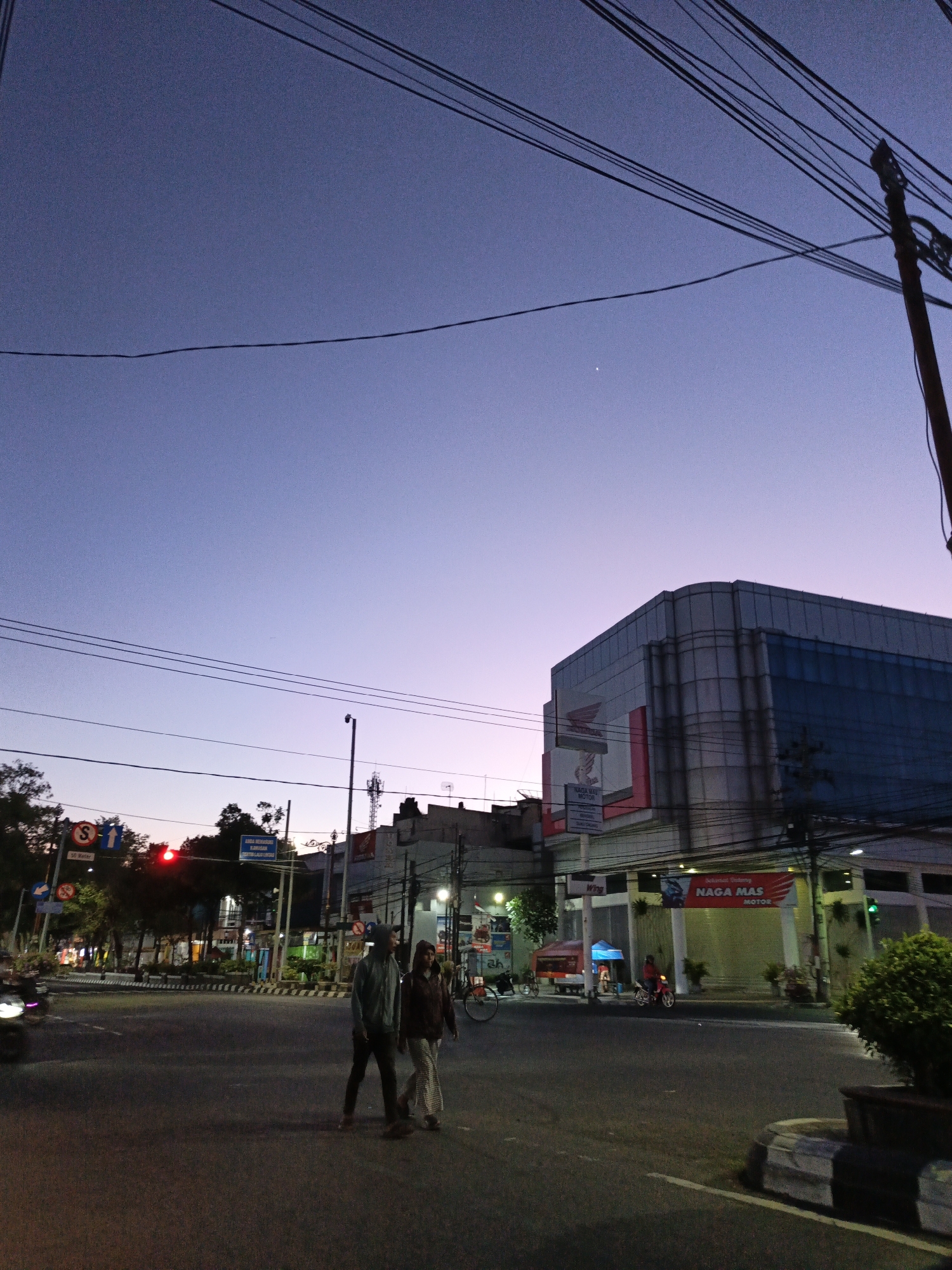 This image depicts a street scene in the city of Klaten, Indonesia, during the twilight hours. The sky is a gradient of deep blue to pale purple, with power lines crisscrossing the frame. On the right side of the image, a modern building with a glass facade dominates the skyline, its architecture characterized by rounded corners and horizontal banding. A prominent sign with red lettering reading "NAGA MAS MOTOR" is visible on the building.
In the foreground, two individuals are crossing a road at an intersection. One person, wearing a hooded jacket, is walking towards the right, while the other, in a lighter colored outfit, walks alongside. The road is paved and appears to be a multi-lane intersection, indicated by lane markings and traffic signals. A red traffic light is visible on a pole to the left. Other elements of the urban environment include trees, streetlights, and a motorcycle with its headlight on in the background. The overall impression is a bustling city street at dusk, with the transition from daylight to evening in progress.