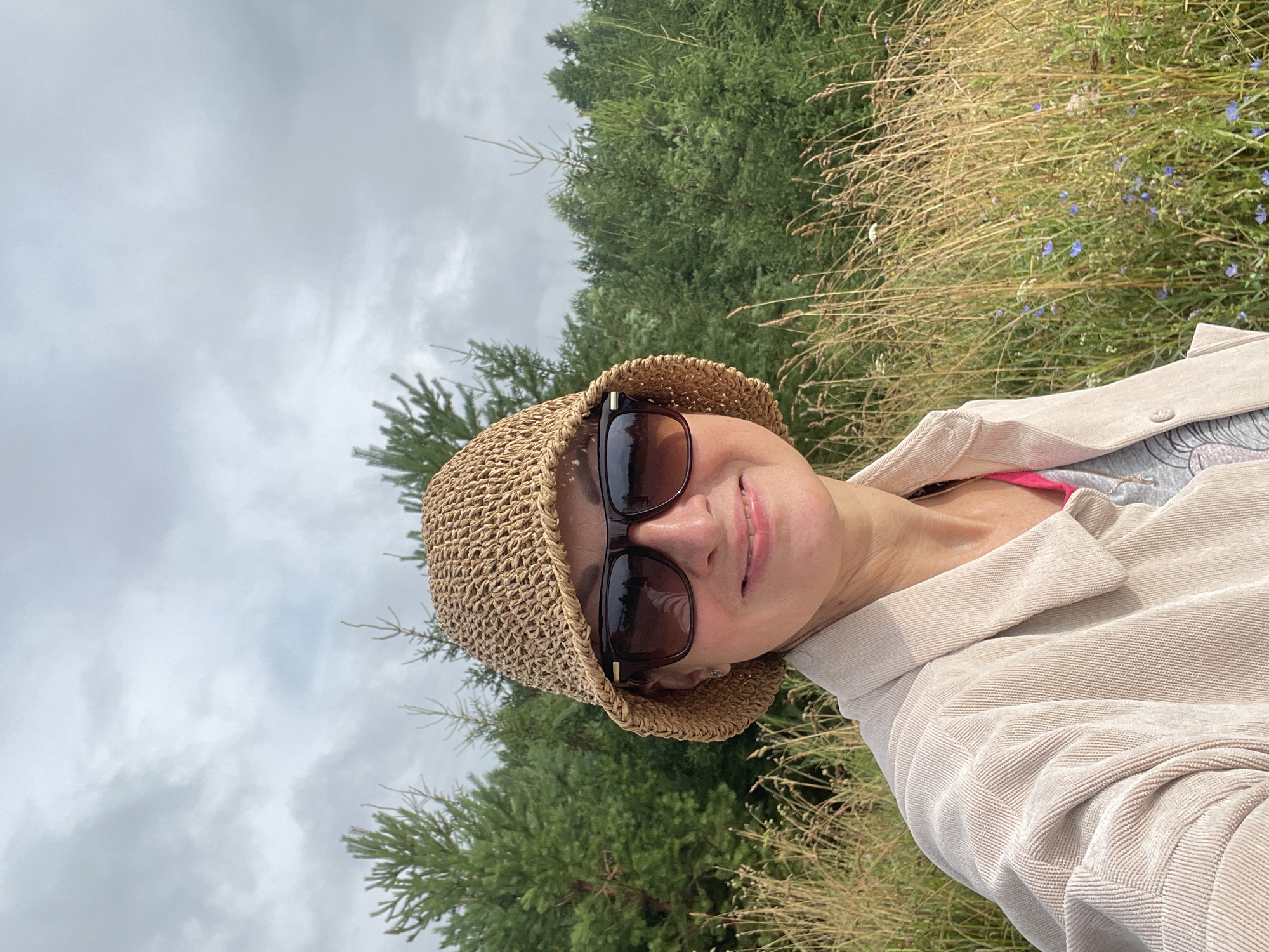 A medium shot shows a woman with fair skin, wearing a straw bucket hat and dark sunglasses. She has on a light-colored corduroy jacket over a pink top and a grey t-shirt. The woman is outdoors, with a background of tall, dry grass, small blue wildflowers, and a dense forest of green pine trees. The sky is overcast with grey clouds. The image is taken from the chest up, with the woman's face slightly turned towards the camera and a smile on her lips. The setting appears to be a natural, rural environment.