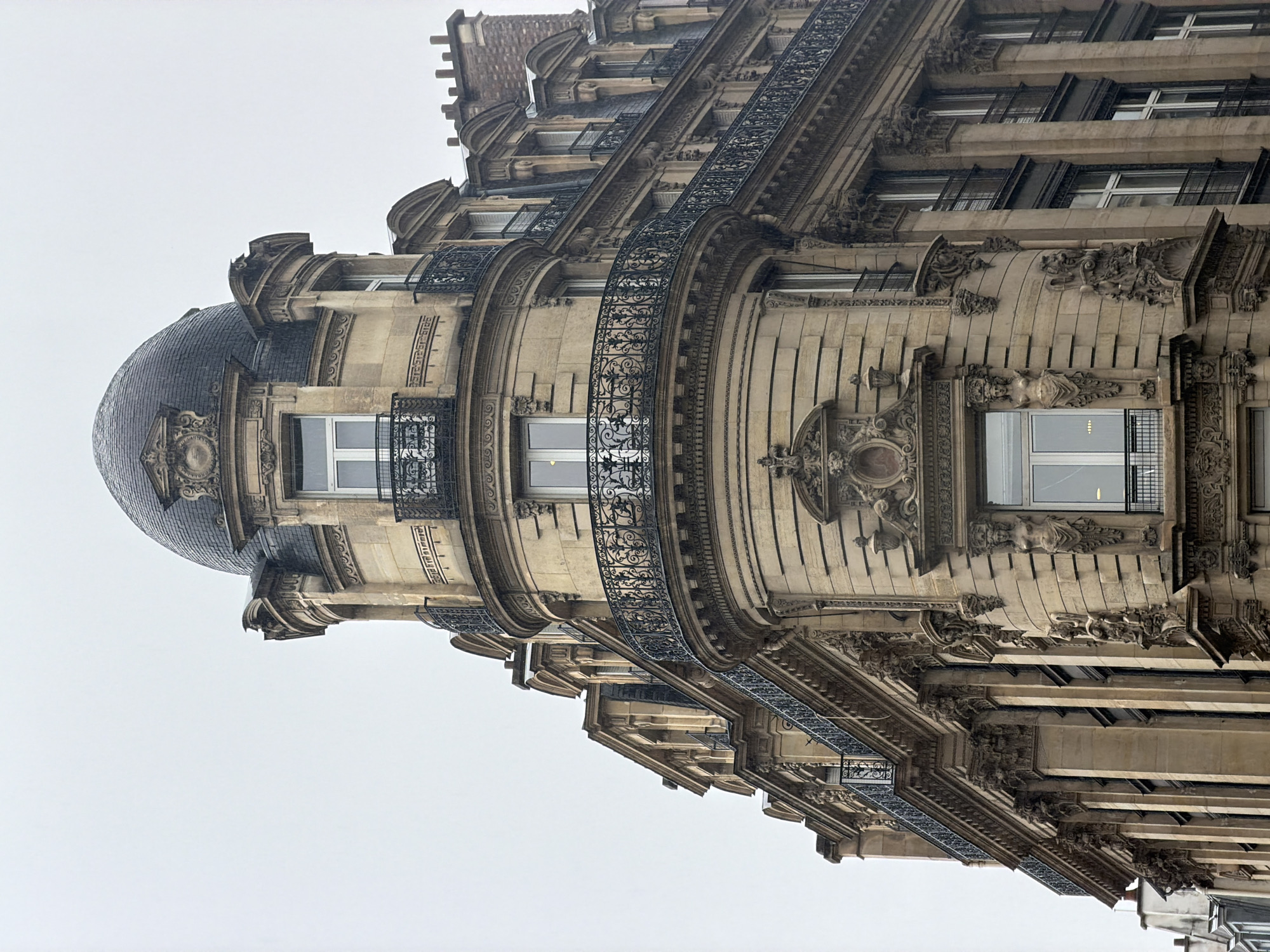 The image depicts the ornate architecture of a building in Paris, France. The building features a rounded corner topped with a dome, balconies with decorative wrought iron railings, and detailed stonework with sculptures and moldings. The windows are rectangular and contain vertical mullions. The sky is overcast and grey.
