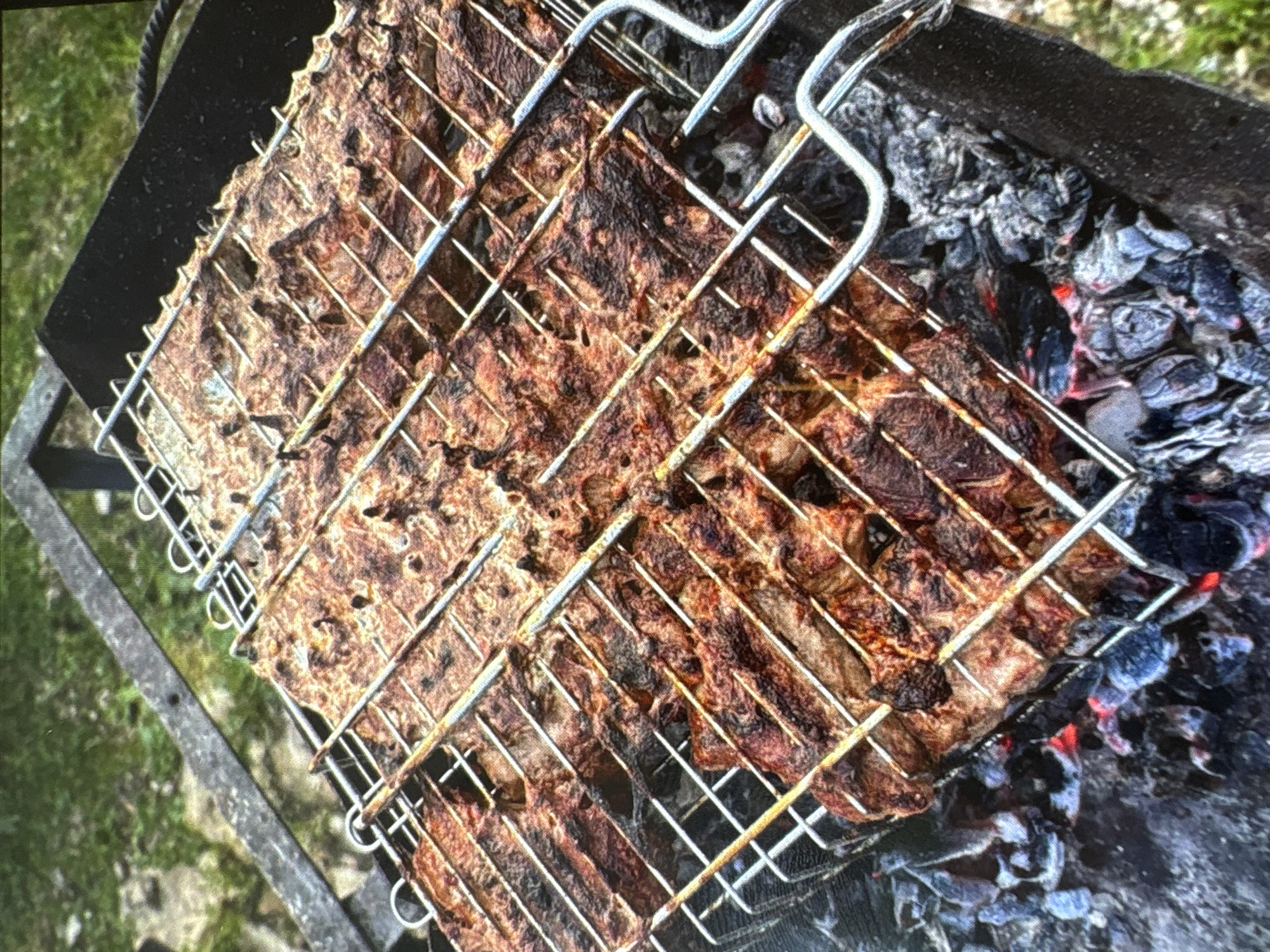 A close-up, high-angle shot shows meat grilling over hot coals. The meat, cut into strips, is arranged on a metal grill basket, which is positioned over a dark-colored barbecue. The coals beneath glow with a reddish-orange hue, indicating intense heat. The background consists of green grass and the edge of a dark metal structure, possibly part of a larger barbecue setup. No people are visible in the image. The scene suggests an outdoor cooking environment. The question about the location in Batumi, Georgia cannot be answered from the provided image.