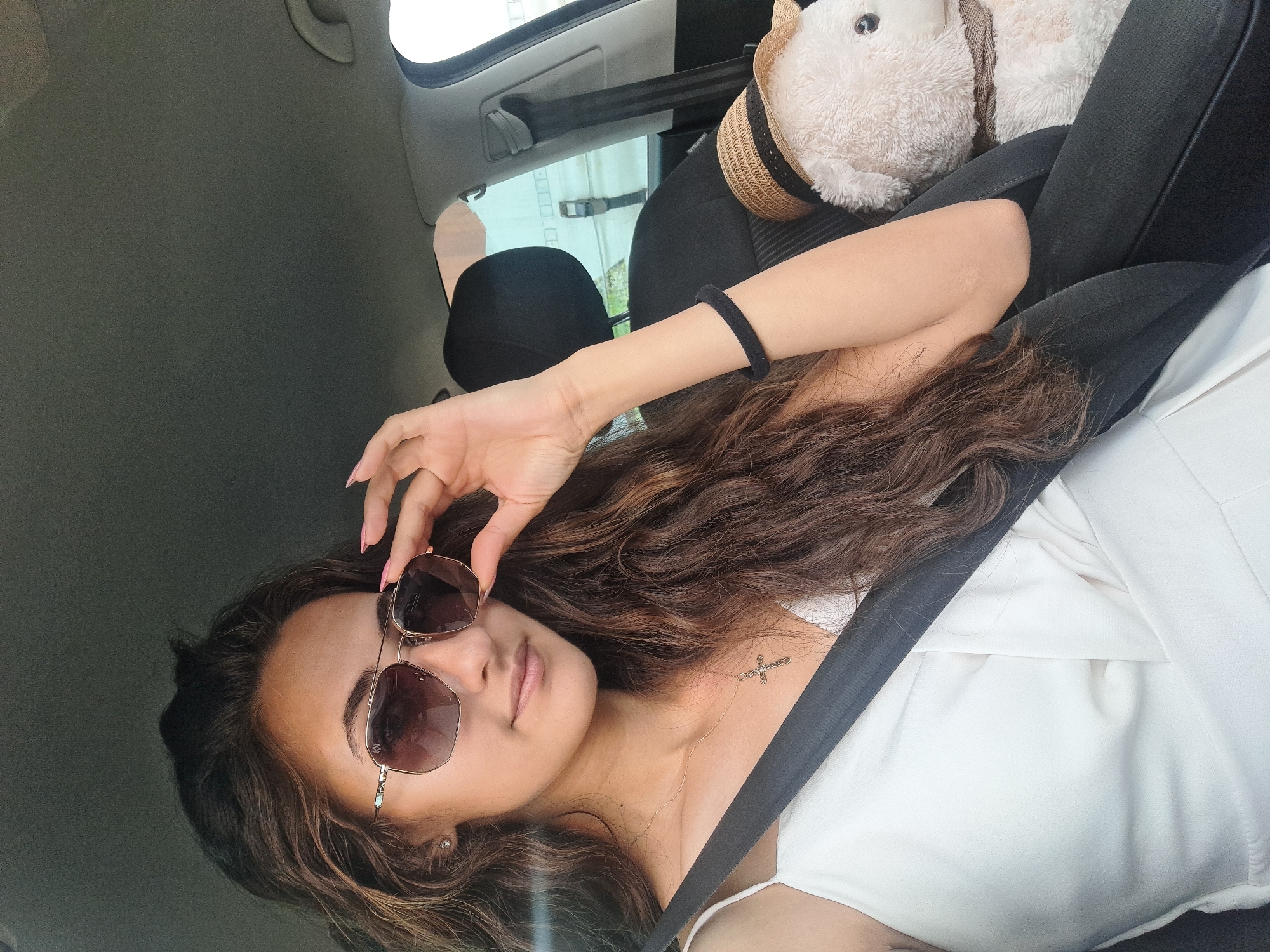 A young woman with long, wavy brown hair and sunglasses is sitting in the passenger seat of a car. She is wearing a white sleeveless top and a gold cross necklace. Her right hand is raised, adjusting her sunglasses. In the backseat next to her, a stuffed teddy bear wearing a straw hat is visible. The car interior is dark, with the seatbelt across the woman's chest. The background shows a bright outdoor scene through the car window.