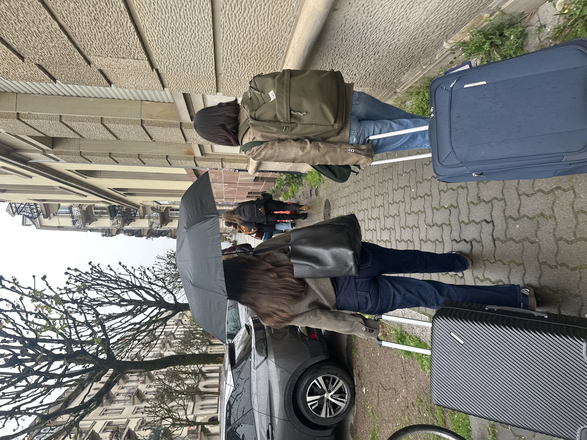 Two individuals are seen walking on a brick-paved sidewalk alongside a street in Strasbourg, France, on a rainy day. The first individual has dark hair, a light brown jacket, blue jeans, and an olive-green backpack. They are pulling a dark blue suitcase. The second individual is holding a black umbrella, has long brown hair, a tan jacket, blue jeans, and a black shoulder bag. They are pulling a black suitcase. A grey car is parked nearby. Other pedestrians can be seen further down the sidewalk. Buildings with multiple floors and windows line the street, with bare trees visible in the foreground. The architecture is consistent with a European urban environment. The scene depicts a typical rainy day in Strasbourg, France, with people navigating the city streets while carrying luggage.