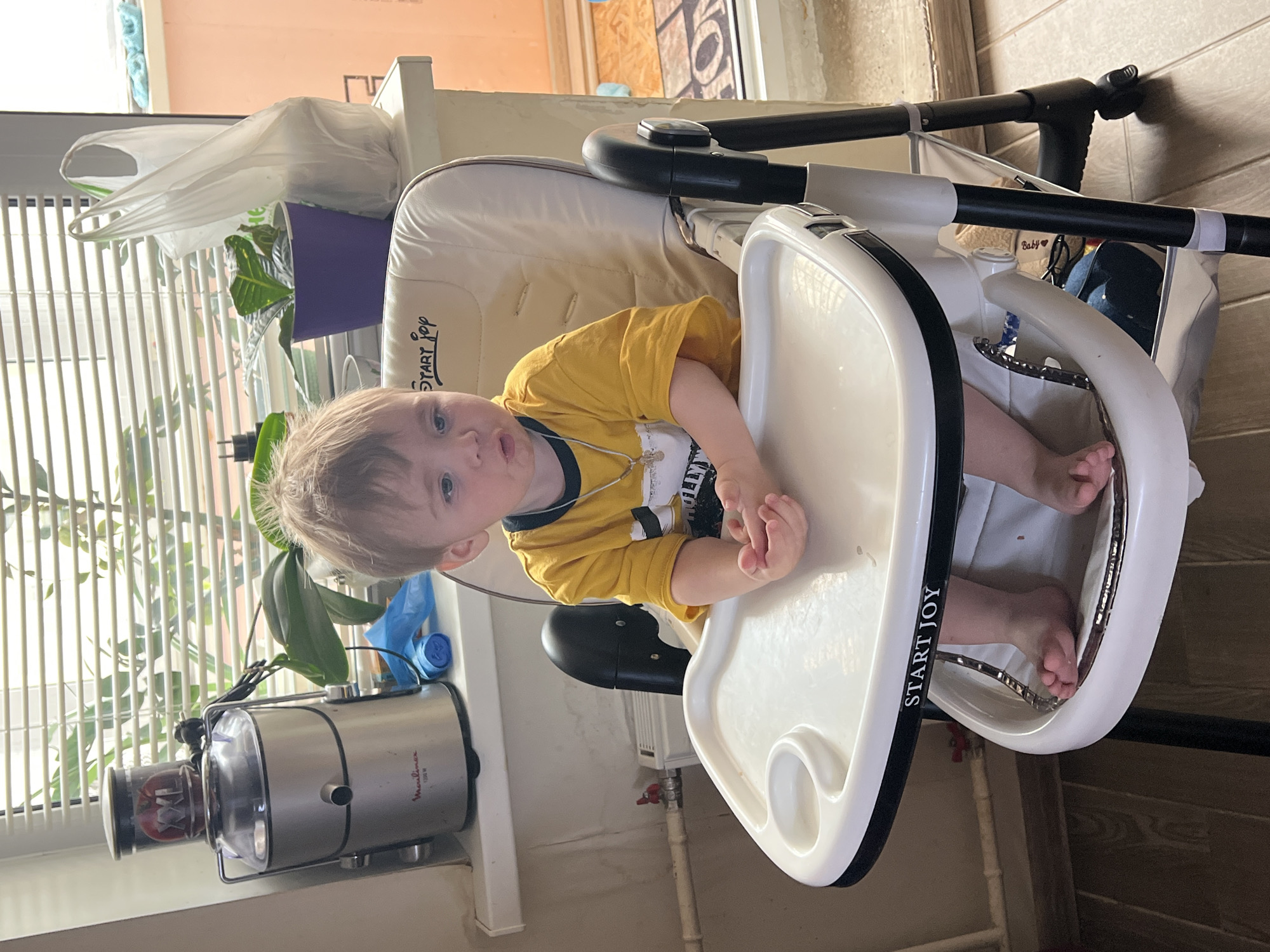 A child is sitting in a white high chair labeled "START JOY". The child is wearing a yellow shirt and has a necklace. The environment includes a Moulinex-branded juicer on a window sill with blinds, a purple plant pot with a plastic bag on top of it, and a tiled floor. The location is in Noviy, Russia, based on the text visible on the item.