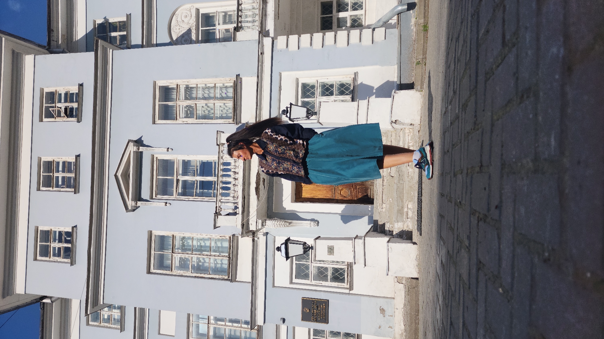 A medium shot captures a woman standing on the steps in front of a pale blue building in Moscow, Russia. The building has numerous windows with white frames and ornate detailing. The woman wears a dark blue jacket with floral embroidery, a teal skirt, and athletic shoes. The background includes a cobblestone street and a drainpipe running down the side of the building.