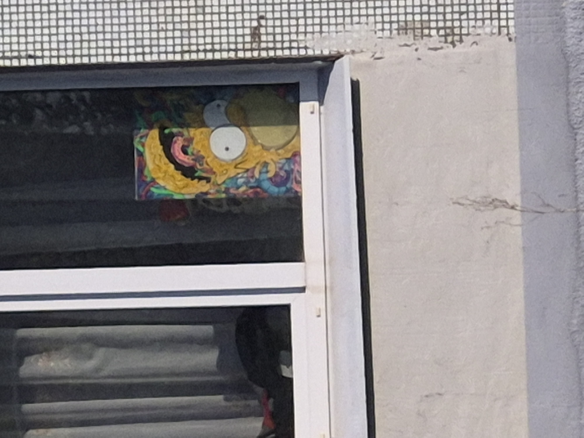 A vibrant, stylized artwork is displayed inside the upper portion of a window, visible from the exterior of a building. The central figure of the artwork strongly resembles Homer Simpson, characterized by yellow skin, large white eyes with small black pupils, and a wide mouth area. However, the rendition is highly abstract and colorful, featuring intricate, swirling patterns in shades of yellow, blue, green, and pink that form what appear to be octopus-like tentacles or an elaborate, psychedelic design around and beneath the character's head.
The artwork is framed by a white window sash. Below the display, the window glass reflects light and shows indistinct interior elements, possibly blinds or curtains. The building's exterior wall to the right of the window is light gray concrete or stucco, exhibiting a rough texture and some minor surface cracks. Above the window, the building facade features a grid pattern of small, light-colored square tiles or mosaic.
The scene is brightly illuminated by natural daylight, indicating it is daytime with clear weather. No specific activity is depicted beyond the static presentation of the artwork. There is no visible text in the image.
