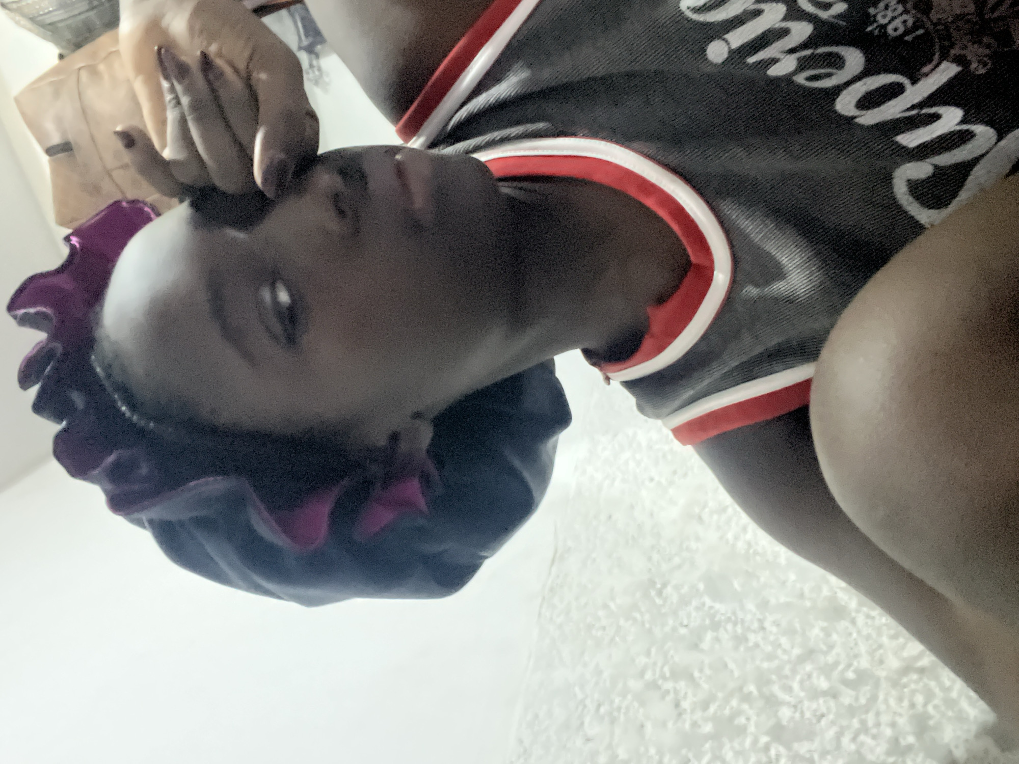 An indoor, close-up, and slightly overhead photograph captures a young person with dark skin, positioned at an angle. The subject is wearing a dark grey or black t-shirt with red and white striped trim around the collar or sleeves. Visible on the shirt is partial white text, appearing to be "Superc" or "Superior" and the year "1985". Their head is covered by a dark purple or black satin bonnet or sleep cap. One hand, adorned with dark nail polish (possibly maroon or dark red), gently rests near their temple, with fingers lightly touching their forehead. The person's eyes are partially visible, looking upwards and slightly to the left from the viewer's perspective, with a neutral or contemplative expression.
The background is simple and light-colored, suggesting an interior space. A textured, possibly tiled or stuccoed, white or off-white surface occupies much of the lower half and right side of the image. In the upper left background, behind the person's hand, there is a blurry light brown or beige object, possibly a bag or item of clothing, contributing to the casual indoor setting. The lighting appears soft and ambient, consistent with an indoor environment. There are no explicit visual cues within the image to indicate the specific location of Abuja, Nigeria, other than the provided context.