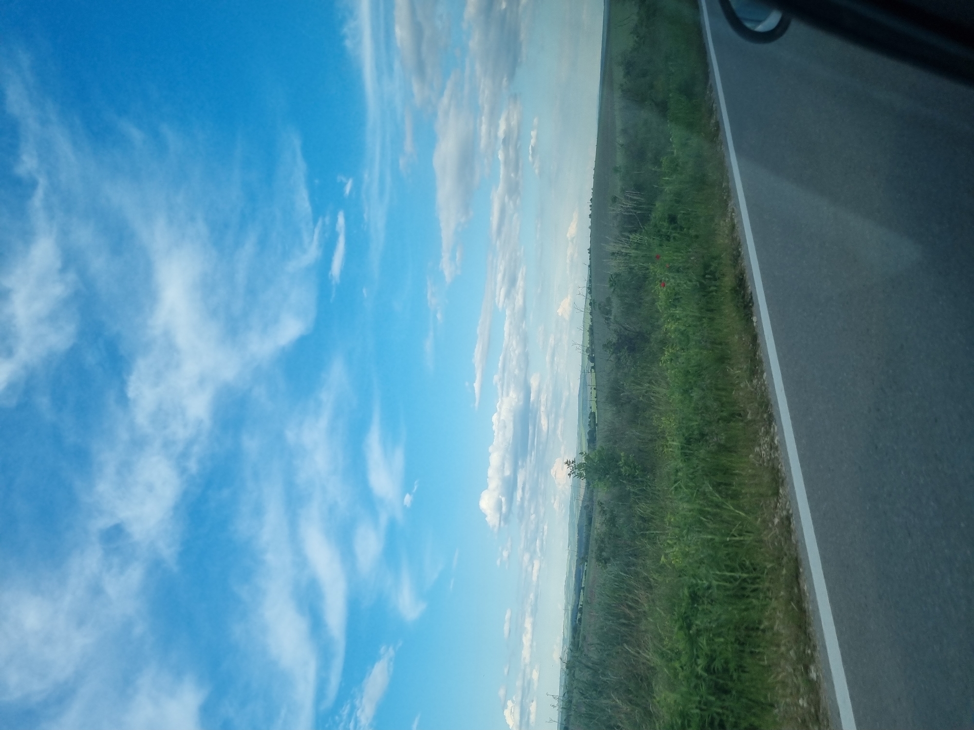 A low-angle shot taken from within a vehicle traveling on a road in Obedinenie, Bulgaria. The perspective is from the passenger side looking towards the sky, a field, and the asphalt road. A bright blue sky occupies the left side of the frame, marked by streaks of cirrus clouds and some cumulus formations near the center, closer to the horizon.
To the right of the sky, a field of lush, green vegetation is visible, with occasional red poppy flowers scattered among the grass. Distant power line structures can be seen on the horizon.
A grey asphalt road with a white dividing line borders the field. The vehicle's window frame is partially visible in the upper right corner. The interior of the vehicle is reflected in the lower portion of the frame, causing a slightly blurred effect on the image.