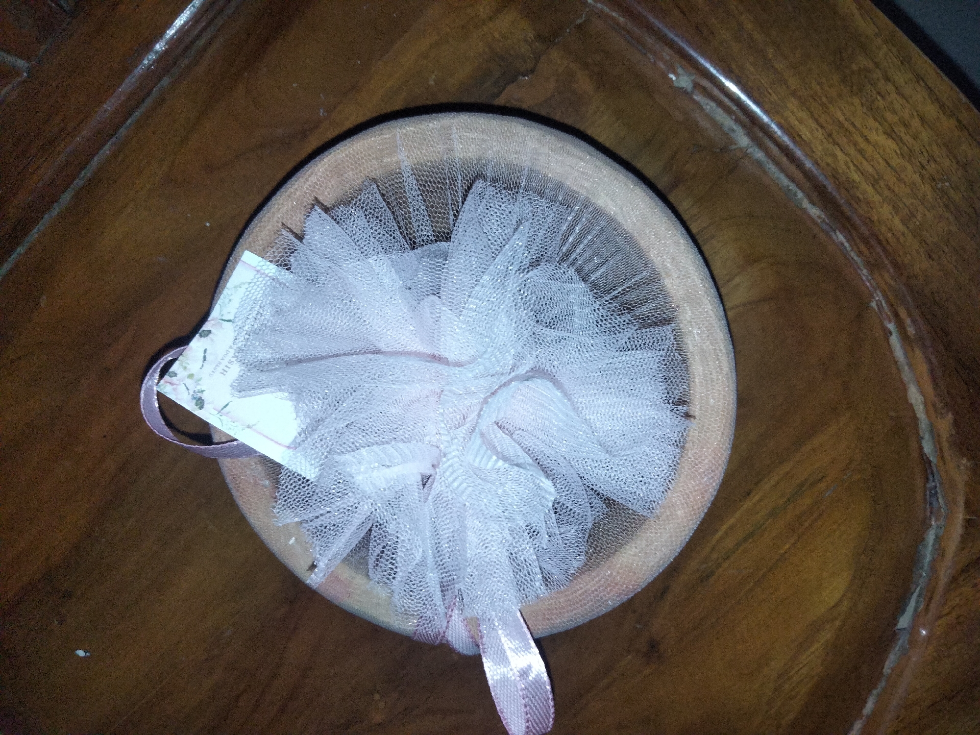 A pale pink mesh bath pouf rests inside a light brown, round bowl. The bowl is positioned on a dark brown wooden surface with a visible grain pattern. A small white tag with some text and floral imagery is attached to the pouf with a pink ribbon. The pouf is fluffy and appears to be made of gathered mesh material.