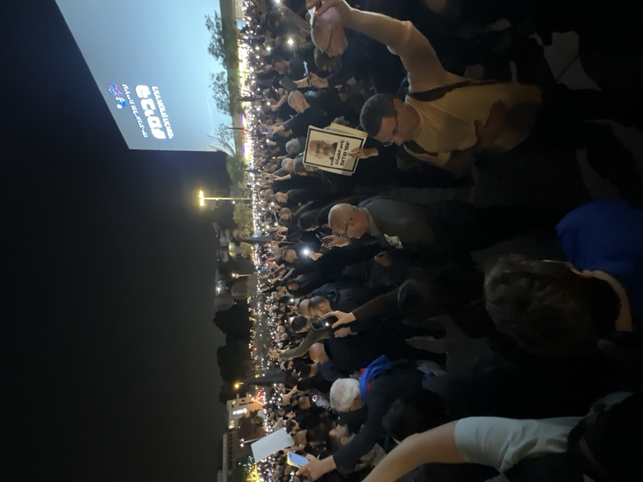The image captures a large outdoor nighttime gathering, with a dense crowd of people filling the frame. The scene is illuminated primarily by numerous small lights held up by individuals in the crowd, which appear to be the flashlights of cell phones, creating a widespread shimmering effect. Bright streetlights and a large projection screen also cast light on the area.
In the upper left portion of the image, a prominent large screen displays text in Hebrew. The top line, in a larger font, reads "מנצחים את החושך" (Menatzchim et HaChoshech), which translates to "Defeating the Darkness" or "We overcome the darkness." Below this, in a smaller font, is "מטה המשפחות להחזרת החטופים והנעדרים" (Mateh HaMishpachot LeHachzarat HaChatufim VeHaNe'edarim), translating to "The Hostages and Missing Families Forum." Further to the left on the screen, below the main text, are the numbers "132" followed by "ימים בשבי" (yamim bashvi), meaning "days in captivity."
Among the crowd, several people are visible holding signs. One sign, held by a person in the mid-ground, features a black-and-white photograph of an older man with glasses and a beard. Below the photo, Hebrew text is written, which reads "זמן לחיות חופשי" (Zman Lichyot Chofshi), meaning "Time to live free." Another plain white sign is visible closer to the foreground.
The activity appears to be a vigil, protest, or demonstration, given the large assembly, the illuminated phones (often used in vigils), and the explicit messages on the screen and signs related to hostages and missing persons. The overall atmosphere conveyed by the illuminated phones and the nature of the messages suggests a moment of solidarity, remembrance, or a plea for action. The multitude of people indicates a significant, well-attended event.