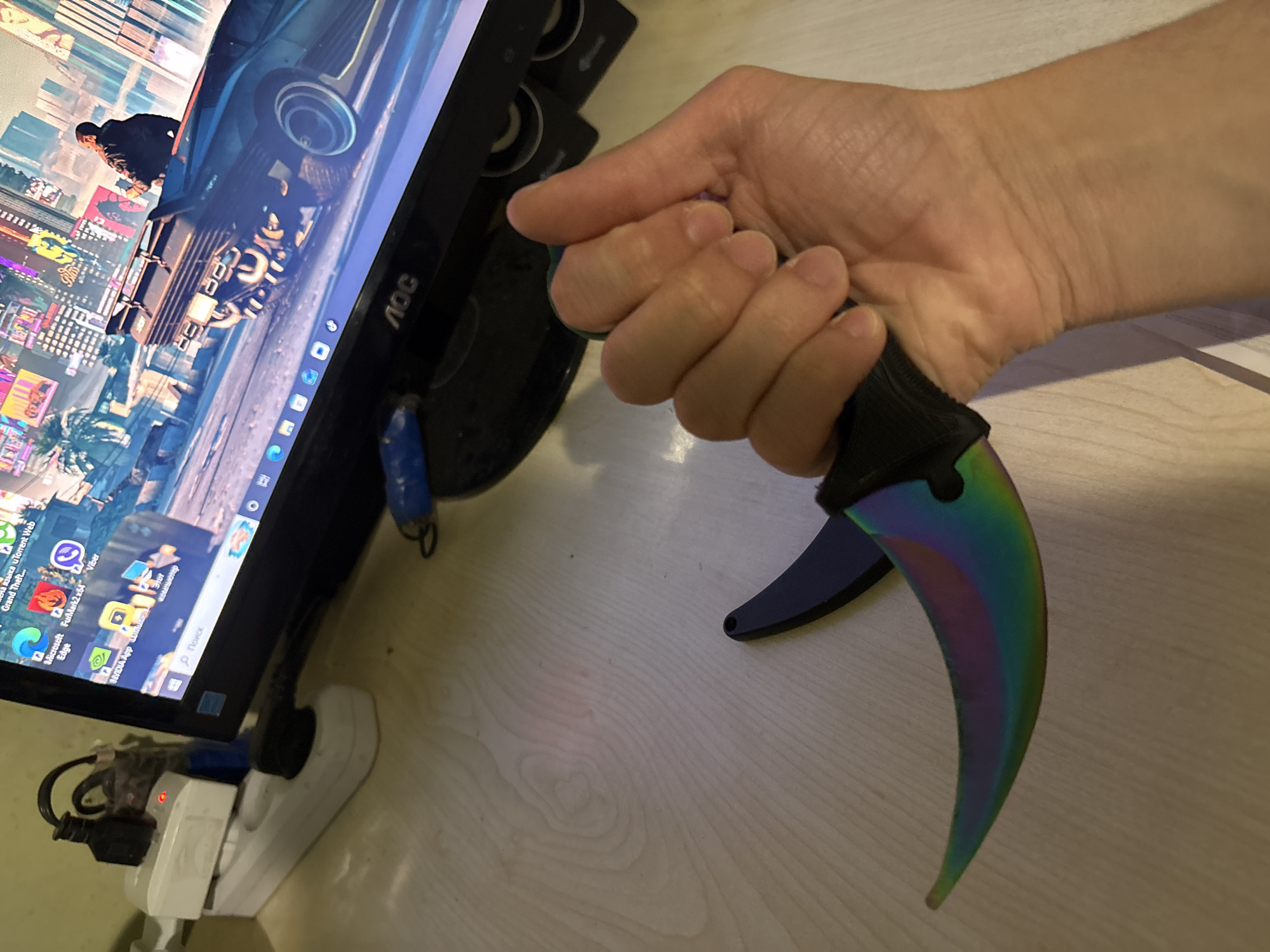 A close-up shot features a hand holding a karambit knife, with an AOC monitor in the background displaying a scene from a video game, possibly Grand Theft Auto. The monitor sits on a desk with an electrical outlet adapter nearby. The scene does not obviously depict Yerevan, Armenia, nor does it depict any people.