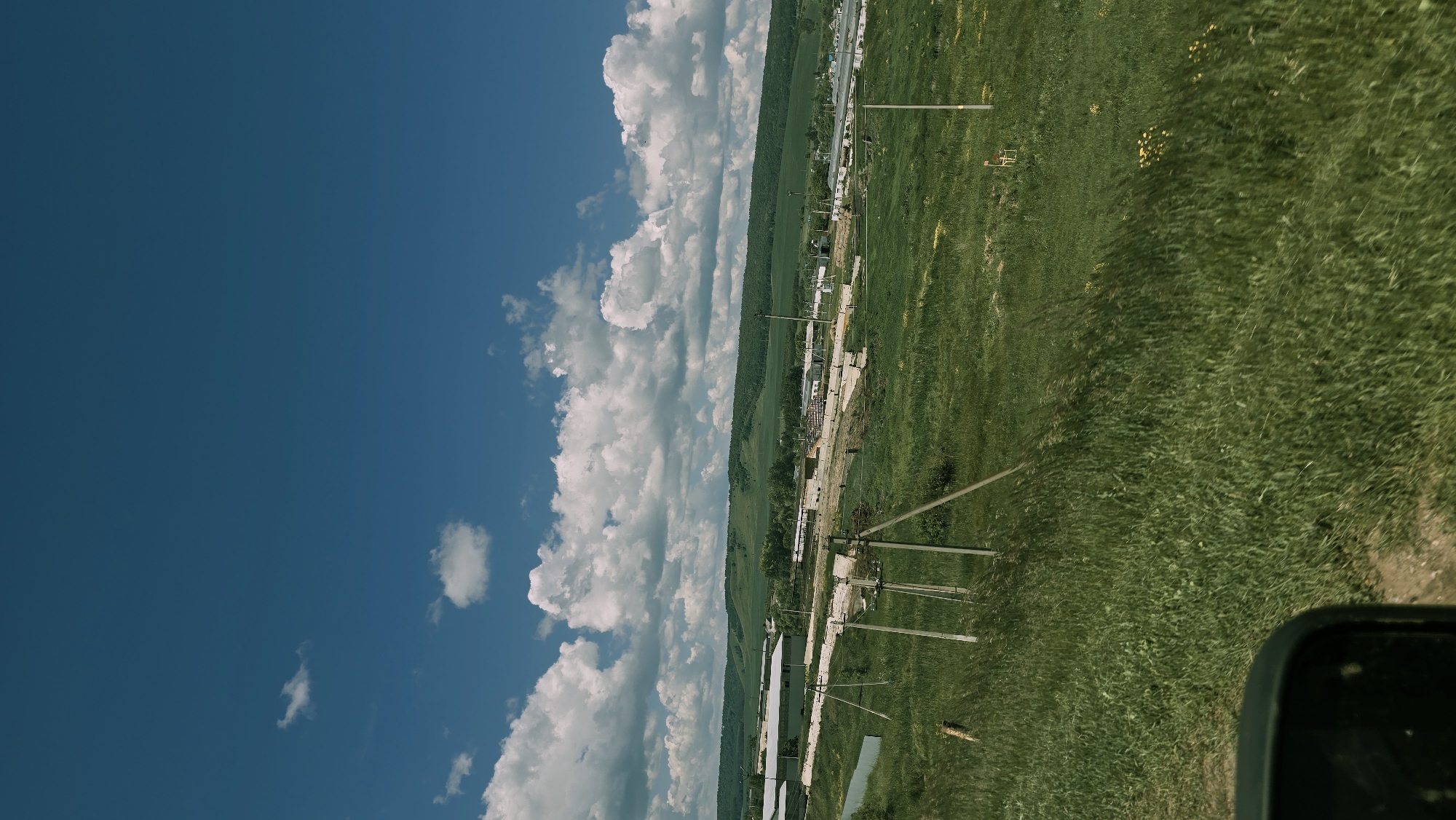 A wide-angle, vertical, medium-shot photograph captures a rural landscape under a bright blue sky with scattered white cumulus clouds. The foreground features a grassy field with tall, green grass. Several wooden poles, likely for power lines, are scattered throughout the field. In the mid-ground, a collection of buildings, possibly agricultural structures or small industrial facilities, are visible. These structures appear to be low-lying and white, with some featuring corrugated metal roofs. Beyond these buildings, a rolling green hillside covered in trees forms the backdrop. The scene suggests a peaceful, agrarian setting within the city of Kudashevo, Russia, under favorable weather conditions. No people are visible in the frame. The composition is dominated by the expansive sky and the natural landscape, with the man-made structures playing a secondary role. The lighting indicates it is daytime, likely late morning or early afternoon, given the bright sunlight and clear sky. The overall impression is one of rural serenity and open space.