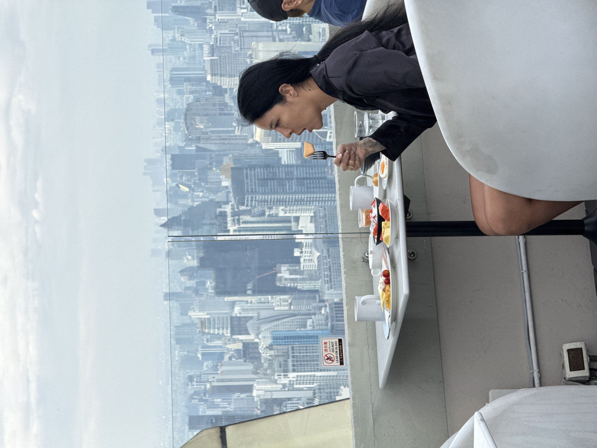 A woman is eating from a plate of food at a table on a high balcony. The background shows an expansive urban landscape, likely the city of Bangkok, Thailand. There are high-rise buildings visible through a glass barrier. The woman is holding a fork with food on it, suggesting she is in the act of eating. The table also has other food items, including a mug and a plate with what appears to be fruit. Another person is slightly visible in the upper left corner. The balcony has a sign that warns against leaning on the edge, and the environment is open-air, high above the city.