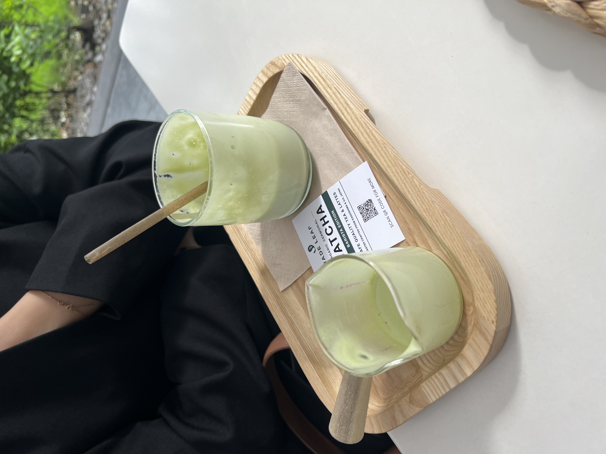 The image shows a person seated at a white table, with a light wood tray holding two glasses of matcha drinks. One glass is nearly full with a wooden straw, while the other glass is partially empty. A napkin and a small card, labeled "JADE LEAF MATCHA," are also on the tray. The person is wearing a black jacket, and a portion of their arm is visible. The background includes greenery, suggesting an outdoor or garden setting.