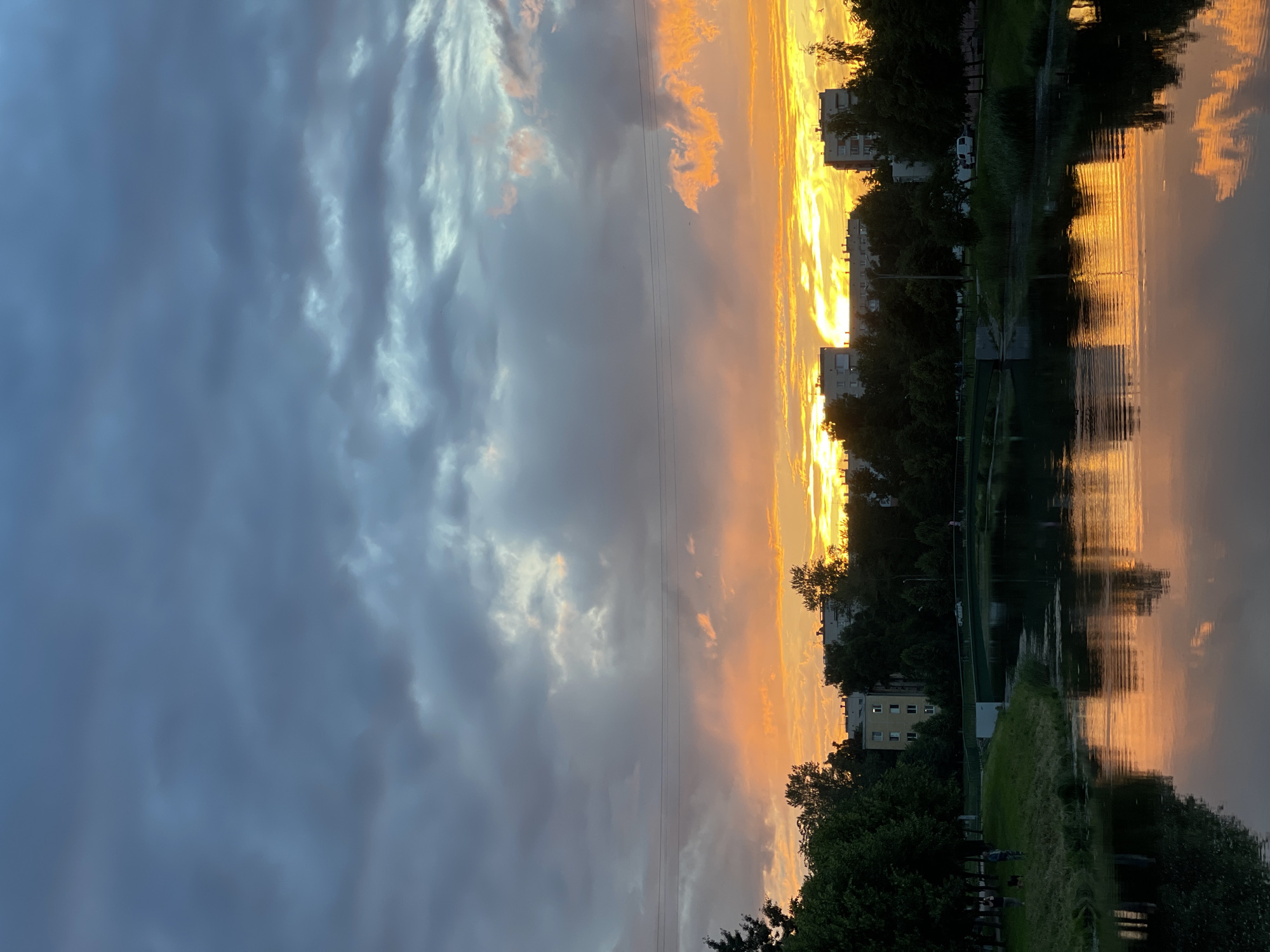 This is a vertical, outdoor, daytime photograph of a sunset over a body of water, likely a canal or river. The sky is dramatic, with dark, moody clouds on the left side and a vibrant band of orange and yellow sunset stretching across the right side, reflected in the calm water. The reflection captures the colors of the sky and the silhouettes of trees and buildings along the opposite bank.
On the bank of the water, a row of trees partially obscures several apartment buildings. The buildings appear to be residential, with multiple windows visible. Some of the trees are tall and full, while others are smaller and more sparse, with visible grass areas. A few people are visible in the distance, sitting or standing on the grassy bank. A white van is parked near one of the buildings. Power lines are visible in the sky.
The time of day is clearly sunset, indicated by the intense colors in the sky and their reflection. The weather appears to be partly cloudy, with both dark and illuminated cloud formations. The scene evokes a sense of peace and natural beauty, contrasted with the urban elements of the buildings. There is no visible text in the image.