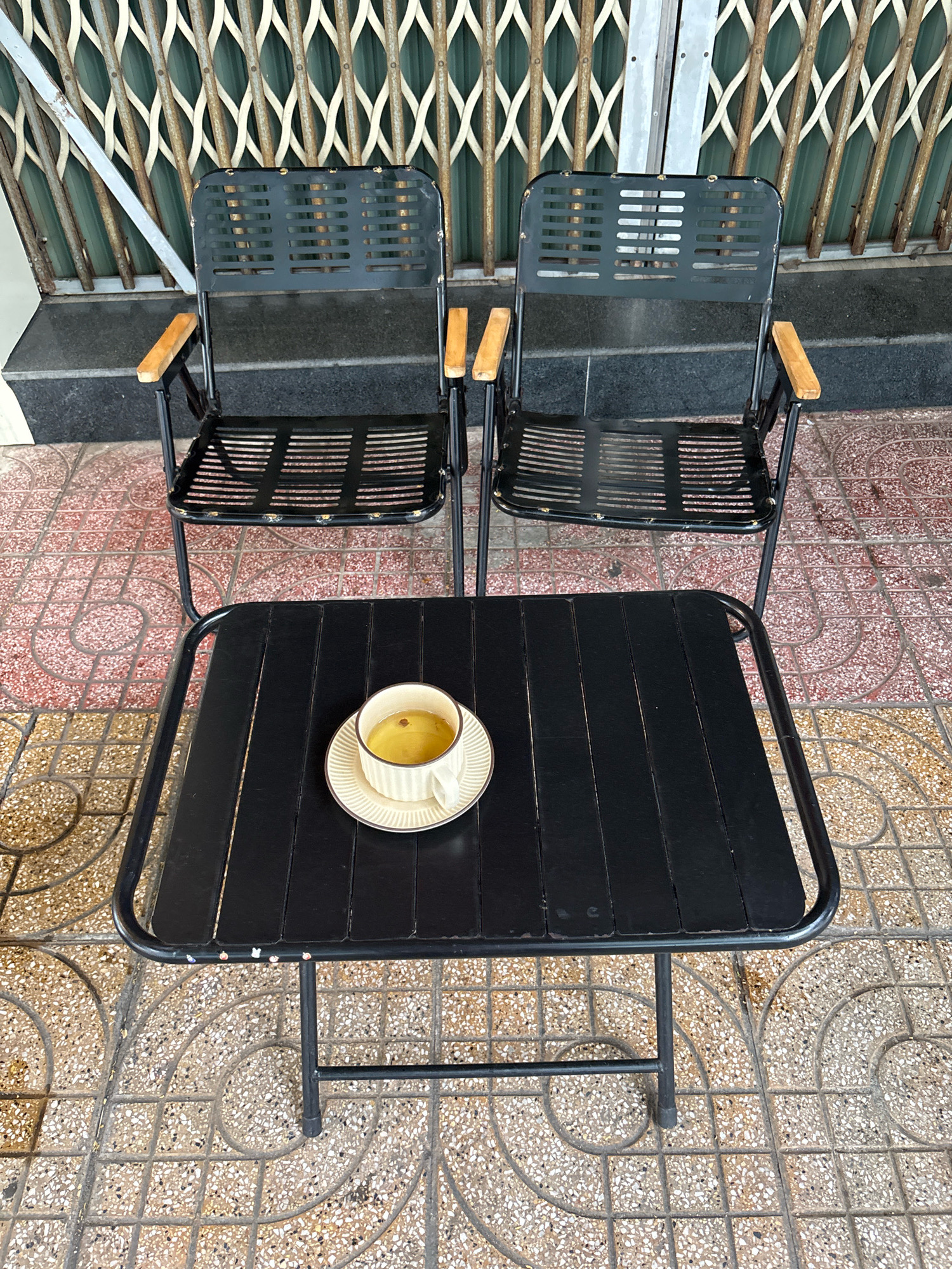 This is a medium-shot, eye-level photograph of an outdoor seating area in Nha Trang, Vietnam. The scene is viewed from a slightly elevated perspective looking down at a black metal table with a cup of light-colored liquid on a saucer, and two black metal chairs with wooden armrests behind it. The table is positioned in the foreground, and the chairs are slightly behind and to the left and right of the table.
The table has a slatted black metal top and thin black metal legs. The cup is white with a subtle pattern around the rim, and it sits on a matching saucer. The liquid inside the cup appears to be a light amber color, possibly tea or coffee.
The chairs are identical, with slatted black metal seats and backs. They have thin black metal frames and thin, rectangular wooden armrests. The chairs are positioned on a patterned tiled floor. The tiles are square and feature a reddish-pink and beige circular pattern.
Behind the chairs, a dark gray, textured surface is visible, possibly a step or base. Further back, a green, metal security gate with a diamond pattern is seen. The gate appears to be attached to a building. The overall environment suggests an outdoor seating area for a business or residence, likely in a commercial or urban setting within Nha Trang. There are no people or animals visible in the image.