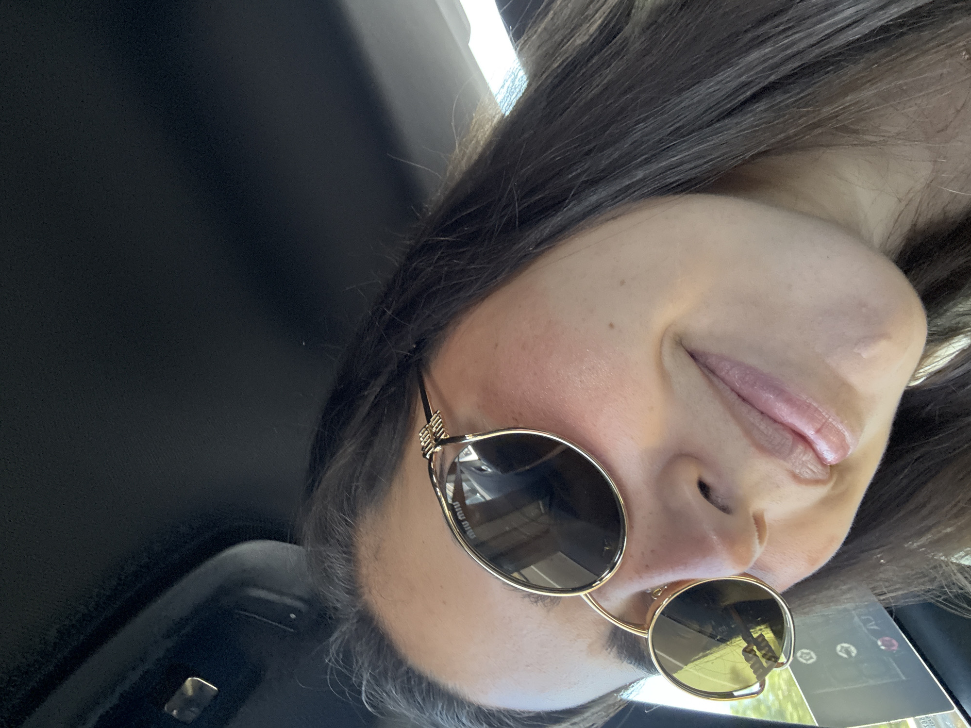 The scene takes place inside a vehicle located in San Matías Tlalancaleca, Mexico. The primary subject is an adult female, shown in a close-up, high-angle view. She has dark brown hair and a slight, closed-lip smile. She is wearing gold-framed, oval-lensed sunglasses with a dark tint; the "Miu Miu" brand name is visible on the left lens. The sunglasses frames feature a decorative gold element at the temples. Her skin exhibits a fair to medium complexion with some redness on the cheeks. The vehicle's dark headliner and an overhead component are visible in the background. Reflections in the sunglasses lenses show an exterior daylight environment, including a partial view of what appears to be a sign or structure displaying white text, possibly "ATJ," against a dark background.