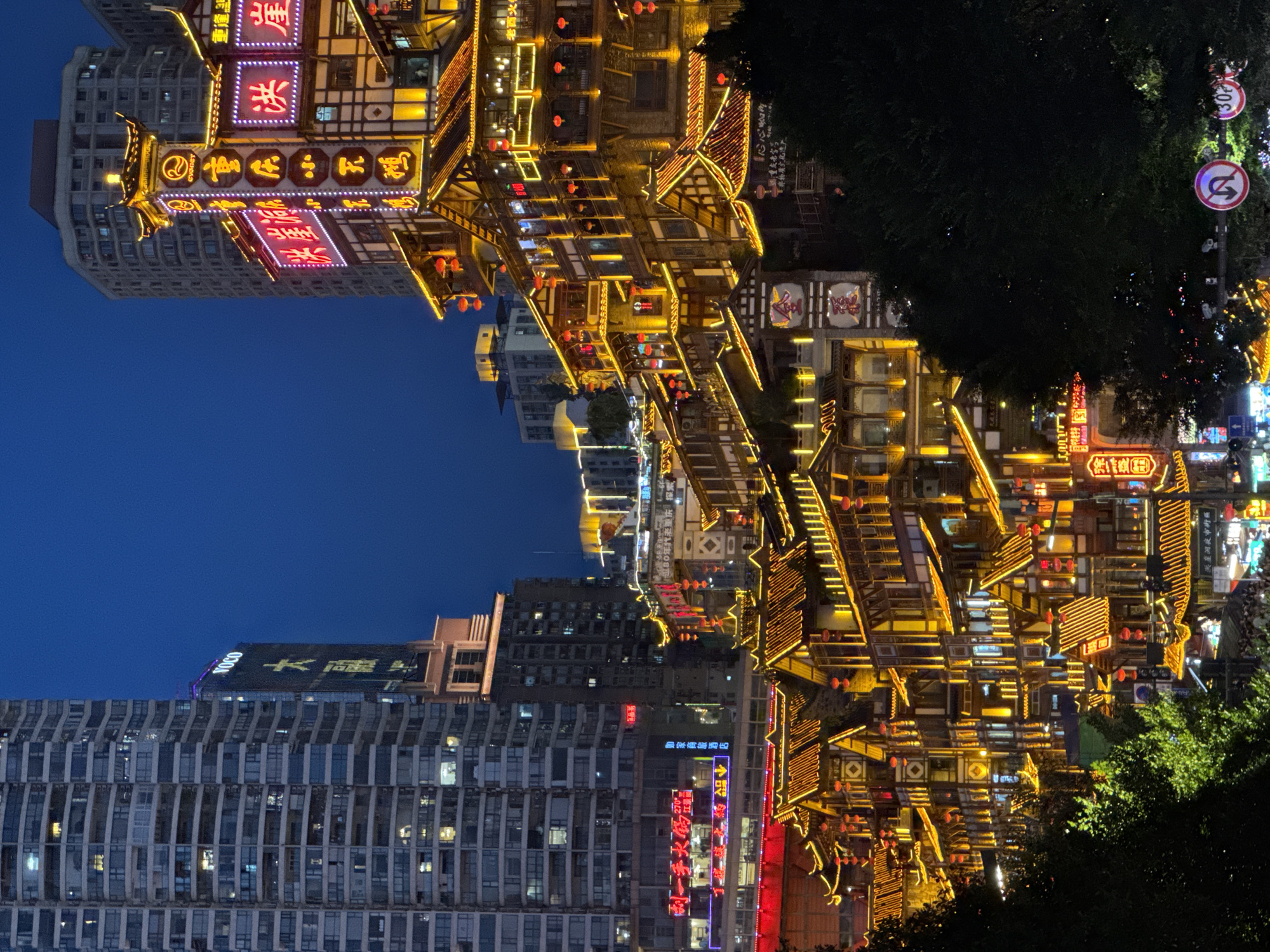 The image captures a vibrant nighttime view of the Hongyadong complex in Yuzhong District, Chongqing, China. The architecture is a distinct blend of traditional stilted buildings adorned with numerous illuminated signs and red lanterns, contrasted against the modern skyscrapers in the background. The scene is dominated by the warm glow of the lights from the buildings, which cascade down the hillside.
In the foreground, dark foliage partially obscures the view, creating a framed effect. The traditional buildings feature multi-tiered structures with intricate wooden detailing and curved roofs, all outlined in bright yellow and orange lights. Several prominent neon signs, in red and white, display Chinese characters, indicating businesses or establishments. Red lanterns hang from eaves and balconies, adding to the festive atmosphere.
Behind the traditional architecture, modern high-rise buildings rise, their windows illuminated, signifying residential or commercial spaces. The sky is a deep, clear blue, indicating it is twilight or night.
On the right side of the frame, there are traffic signs. One circular sign with a red border shows a black arrow indicating a U-turn is prohibited. Another sign, also circular with a red border, displays the number "30" in black, likely indicating a speed limit.
Below the main Hongyadong structures, hints of a bustling street scene are visible with more lights and what appear to be people walking. The overall impression is one of a dynamic urban landscape where historical architecture meets contemporary development, all bathed in the captivating light of the night.