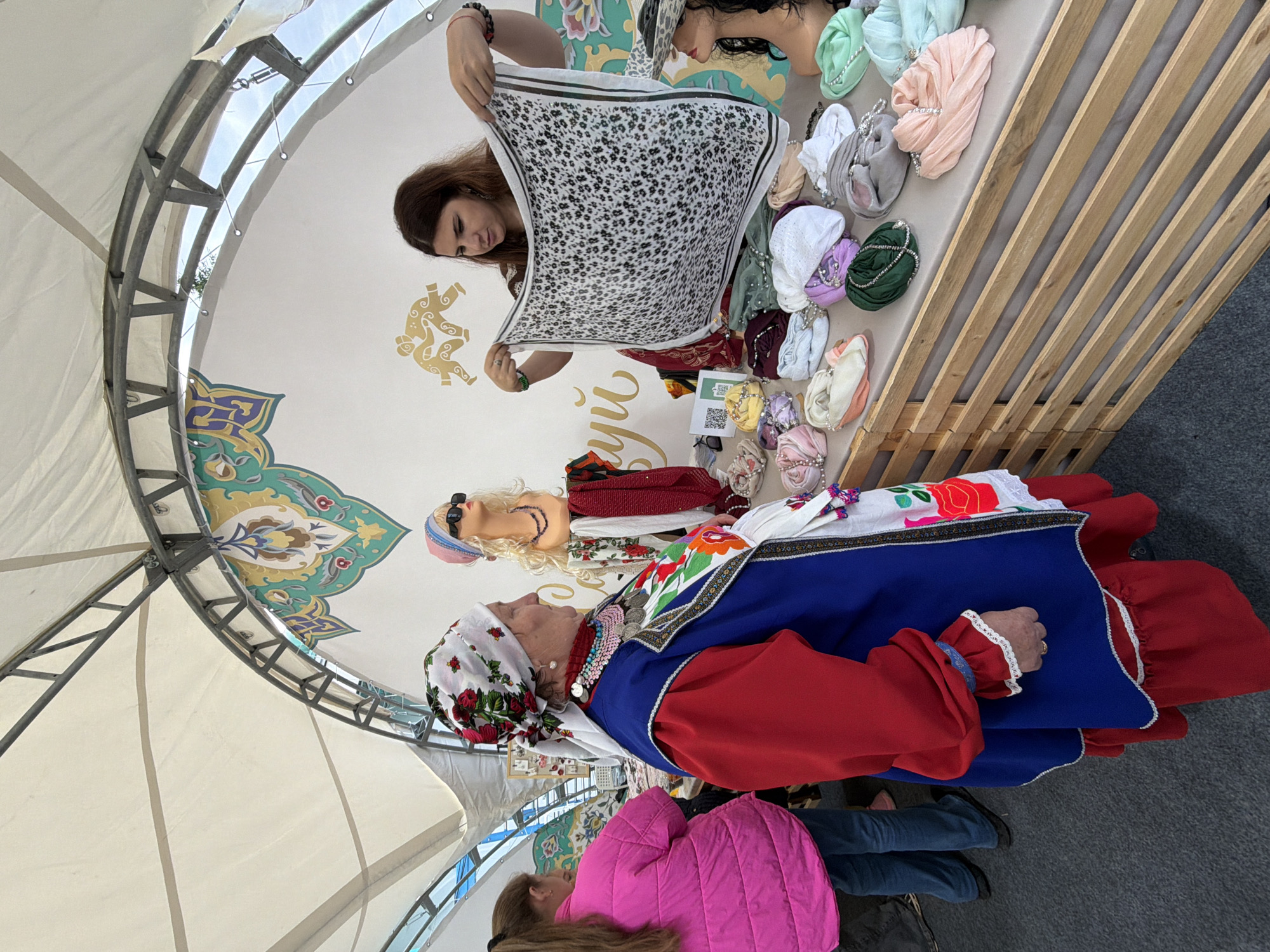 A woman is displaying a patterned scarf at an outdoor market stall. She holds the scarf with both hands, showing its design to an older woman dressed in traditional clothing. The stall features a variety of colorful scarves and headwear displayed on a wooden table. Behind the stall, mannequins are adorned with headscarves and sunglasses. The market appears to be set up under a white canopy supported by metal beams. The backdrop includes decorative wall art with symbolic imagery and some text.