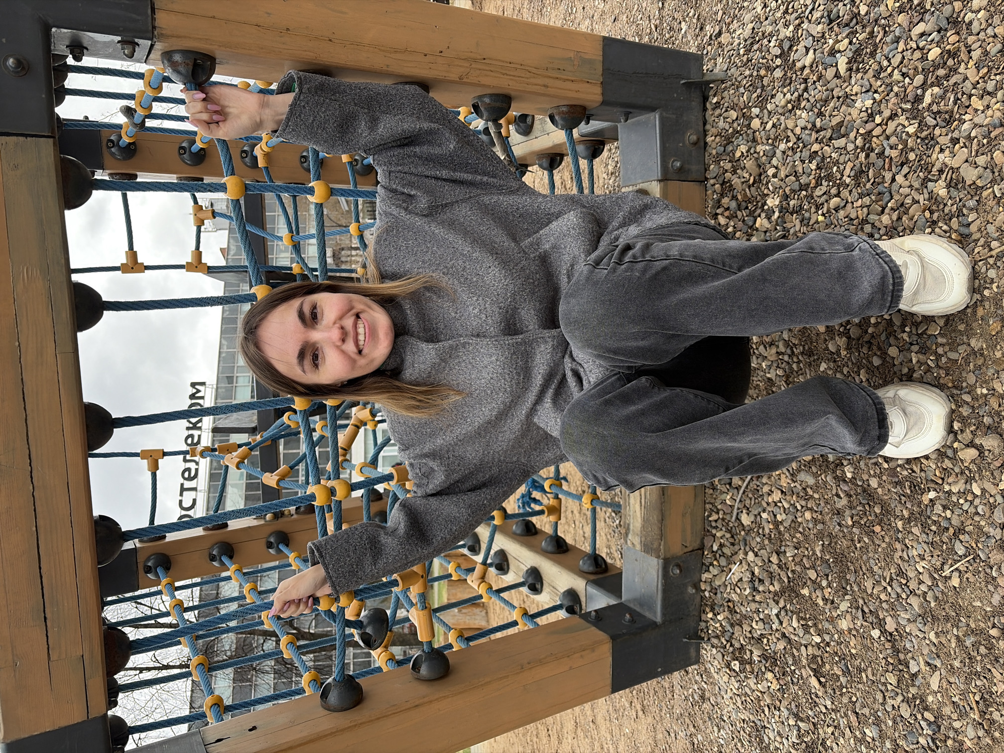 A young woman is captured in a full-body, smiling portrait at an outdoor playground in Izhevsk, Russia. She is positioned on a climbing structure, leaning against or sitting on a part of it, holding onto blue ropes and black grips with both hands. Her face is turned towards the camera, beaming with a wide, cheerful smile.
She is dressed in casual attire, featuring a grey, textured turtleneck sweater or coat and dark grey, possibly faded black, loose-fitting jeans. Her feet are visible, wearing clean white sneakers with a thick sole, resting on the gravel ground. The climbing structure is made of sturdy wooden beams, dark metal supports, and an interconnected network of blue ropes punctuated by yellow and black plastic or rubber connectors and grips.
The setting is outdoors, likely during the day, under an overcast sky which provides soft, diffused light. The ground is covered with light-colored gravel. In the background, a multi-story building with numerous windows is visible, and prominent text "ОСТЕЛЕКОМ" (Rostelecom), a Russian telecommunications company, can be clearly read on its facade, indicating an urban environment. The image conveys a lighthearted and joyful mood, with the woman appearing to enjoy her time at the playground.