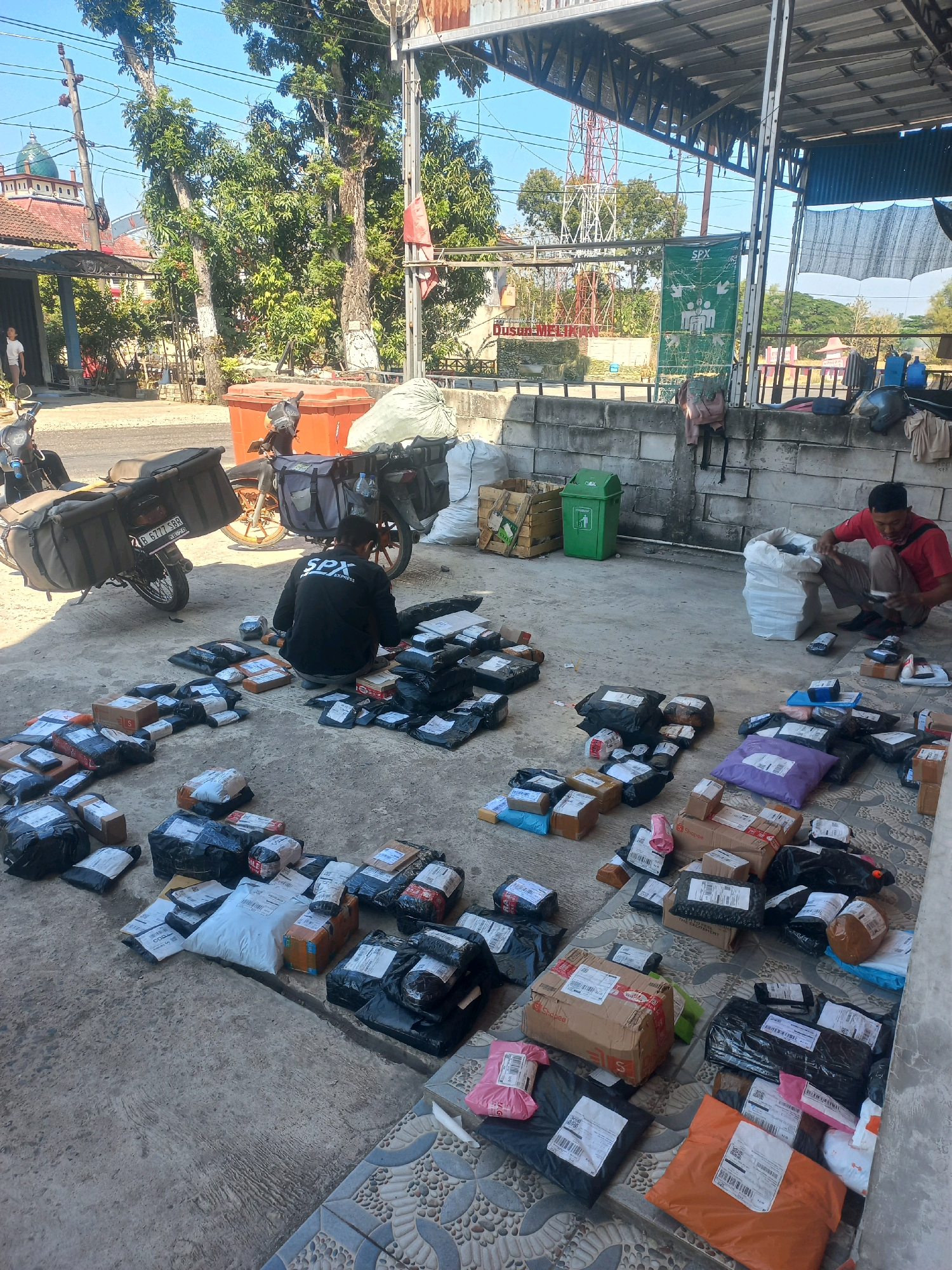 In Baturetno, Indonesia, on a sunny day, a man wearing a black shirt with "SPX Express" branding sits on the ground, sorting through a large pile of packages. The packages, varying in size and packaging, are spread across a concrete floor. Another man in a red shirt crouches nearby, also interacting with a large white bag of packages. Two motorcycles, one with a large cargo box and the other with a smaller one, are parked behind the men. In the background, a brick wall borders the area, and a green trash can is visible. Beyond the wall, trees and a building with a green dome suggest a semi-rural or suburban setting. The overall scene depicts a logistics or delivery operation in progress.