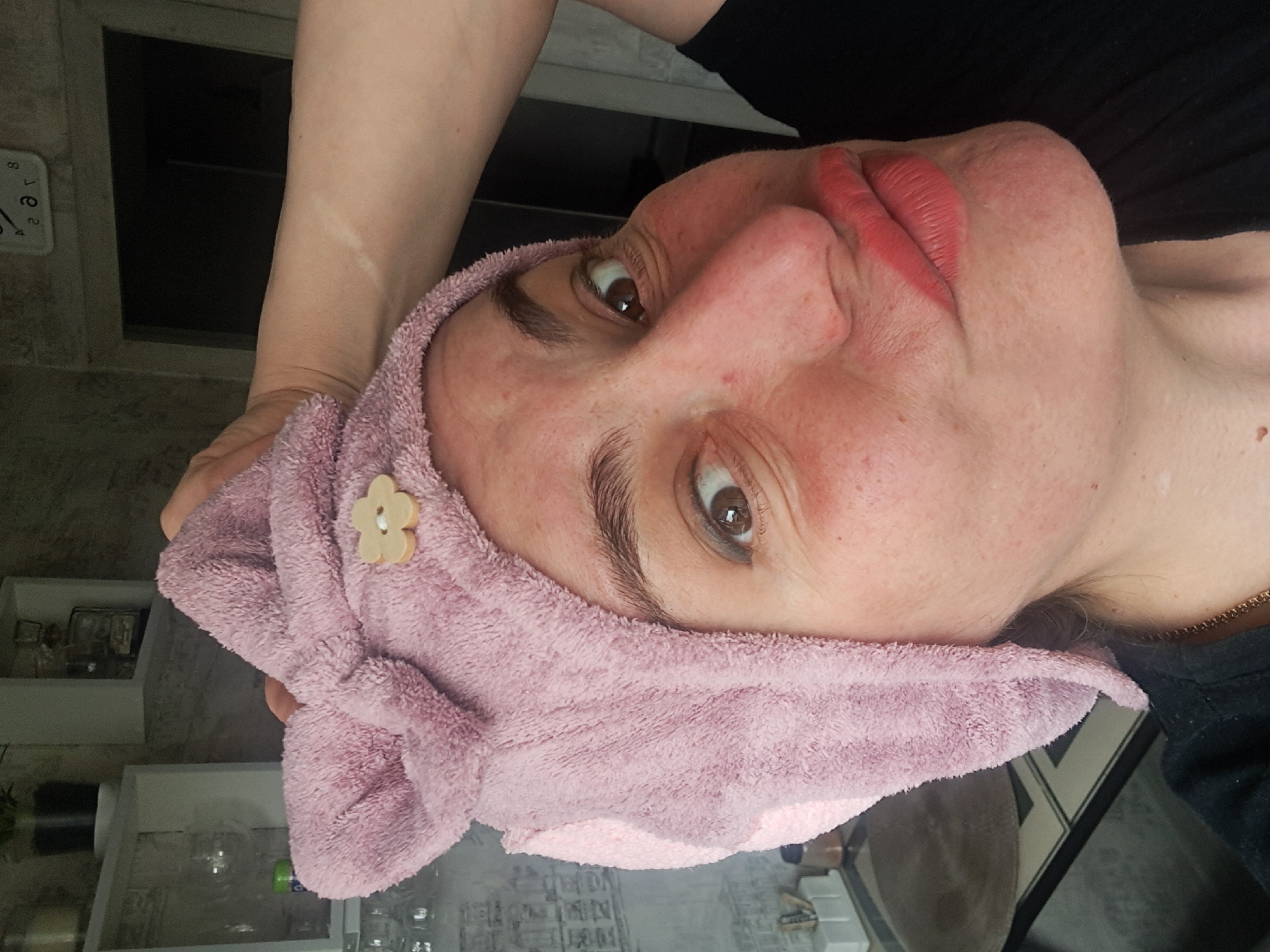 A close-up, slightly rotated image depicts a woman with a pink towel wrapped around her head, adorned with a light-colored flower-shaped ornament. She has reddish skin, brown eyes, and red lipstick. The background shows a room interior with shelves, glass objects, and possibly a mirror. The location in the city of Elektrostal, Russia, cannot be definitively determined from the image.