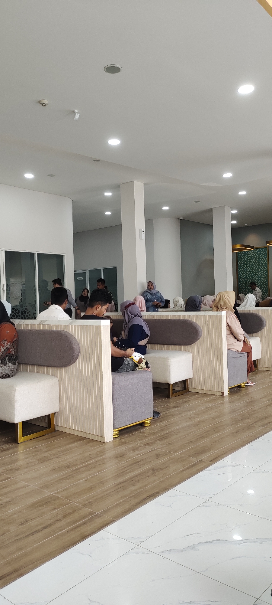 The image captures a bright, modern indoor waiting area, likely located in Lhoksukon, Indonesia. Several individuals are present, seated in comfortable, contemporary armchairs and benches arranged in rows, separated by low, paneled dividers.
In the foreground and midground, multiple people are seen. A man in a black t-shirt sits with his head bowed, seemingly focused on something in his lap. Beside him, a woman wearing a purple hijab and a dark top is also seated. Further back, another woman in a light brown hijab sits casually. Many of the women in the scene are wearing hijabs, indicating a cultural setting common in Indonesia. Other individuals are visible in the background, some standing near glass partitions and others seated further back.
The interior design features white walls and a white ceiling equipped with numerous recessed circular lights, providing bright illumination. A circular speaker and a smoke detector are also mounted on the ceiling. The seating units are upholstered in light grey and off-white fabrics, with some having golden-colored metal bases. The dividers between the seats are light-colored with vertical paneling. The flooring transitions distinctly from a dark wood-look tile to a lighter, white marble-patterned tile, diagonally crossing the space. In the background, a decorative screen with an intricate geometric pattern adds visual interest. The atmosphere appears calm and orderly, with people engaged in waiting or quiet personal activities. There is no visible text in the image.