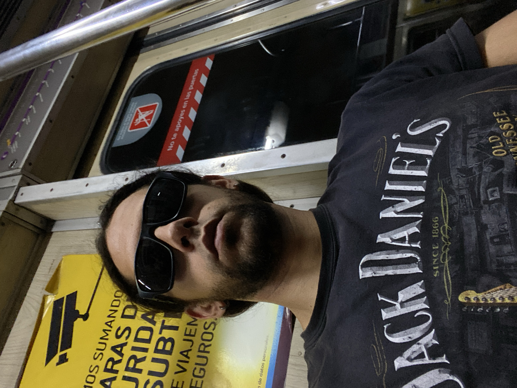 The image is a selfie of a man wearing sunglasses and a Jack Daniel's t-shirt on what appears to be a subway car in Buenos Aires, Argentina. A sign on the subway car reads "No te apoyes en las puertas", which translates to "Do not lean on the doors". Another sign indicates that CCTV is used for security.