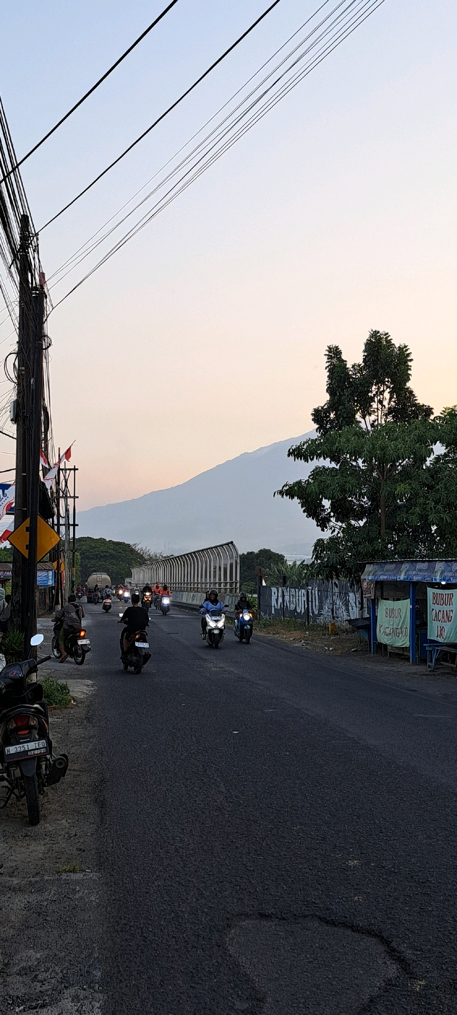 This image captures a street scene in Randupitu, Indonesia, at dusk. The sky is a soft gradient of pale blue and peach, with numerous power lines crisscrossing the frame. A road runs diagonally through the scene, populated by several motorcycles carrying riders. Some motorcycles are parked on the side of the road, including one in the foreground with a license plate reading "H 3351 TEQ".
On the left side of the road, a utility pole stands tall, adorned with several Indonesian flags and various signs. To the right of the road, there's a small roadside stall with blue awnings and signs that appear to advertise food, with "BUBUR CACANG" visible. A large tree with dense foliage stands behind the stall. In the background, a massive, hazy mountain range dominates the horizon, with a long, white-railed overpass crossing in front of it. The road itself shows signs of wear, with patches and cracks visible. The overall impression is a typical evening commute in a semi-rural Indonesian setting.