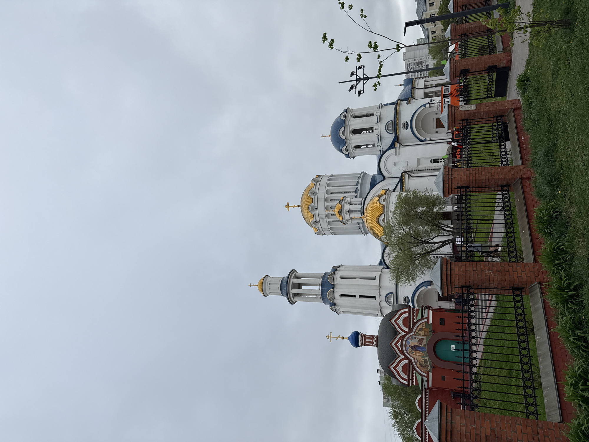 The image depicts an Orthodox church complex in Moscow, Russia, under a cloudy sky. The buildings consist of white structures with blue and gold domes, typical of Russian Orthodox architecture. The church is surrounded by a brick and wrought-iron fence, and there is greenery along the edge of the property. A small, red chapel with religious artwork adorns the front of the complex. The scene appears to be located within an urban setting, as other buildings can be seen in the background. The general impression is one of a peaceful, historical landmark within a modern city.