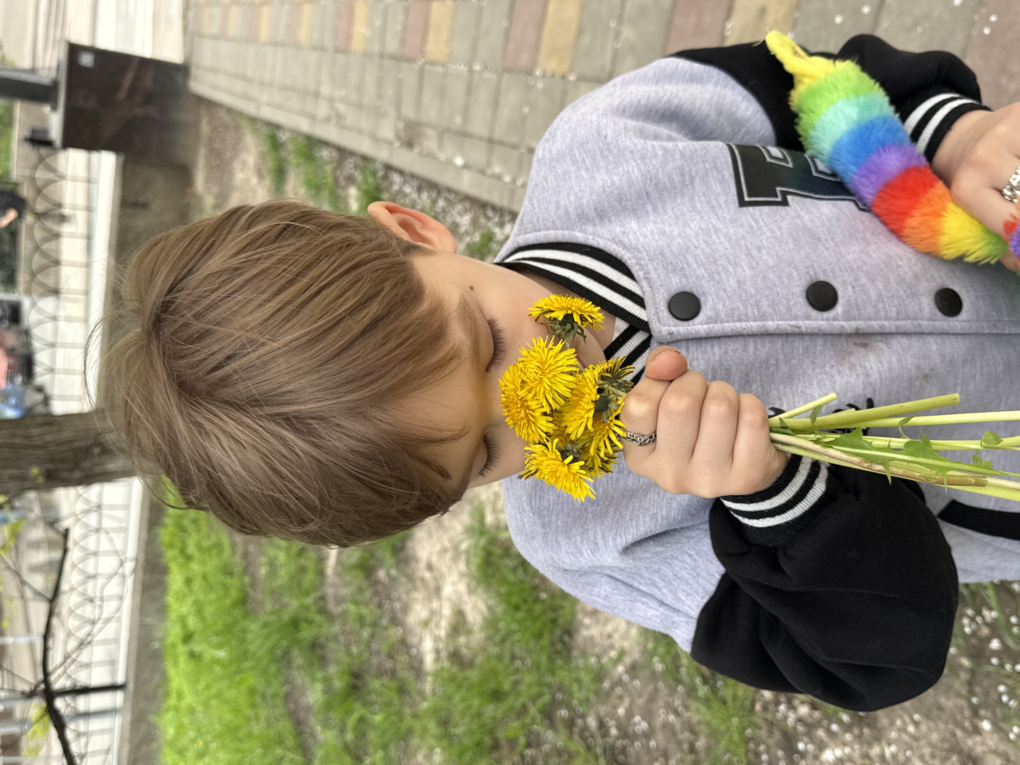 A child with light brown hair is holding a bouquet of yellow dandelions to their face, appearing to smell them. The child is wearing a gray and black letterman-style jacket with a black and white striped collar. They also have a rainbow-colored, fuzzy object in their hand. The background shows a paved sidewalk and some green grass, suggesting an outdoor location. The scene takes place in Rostov-on-Don, Russia.