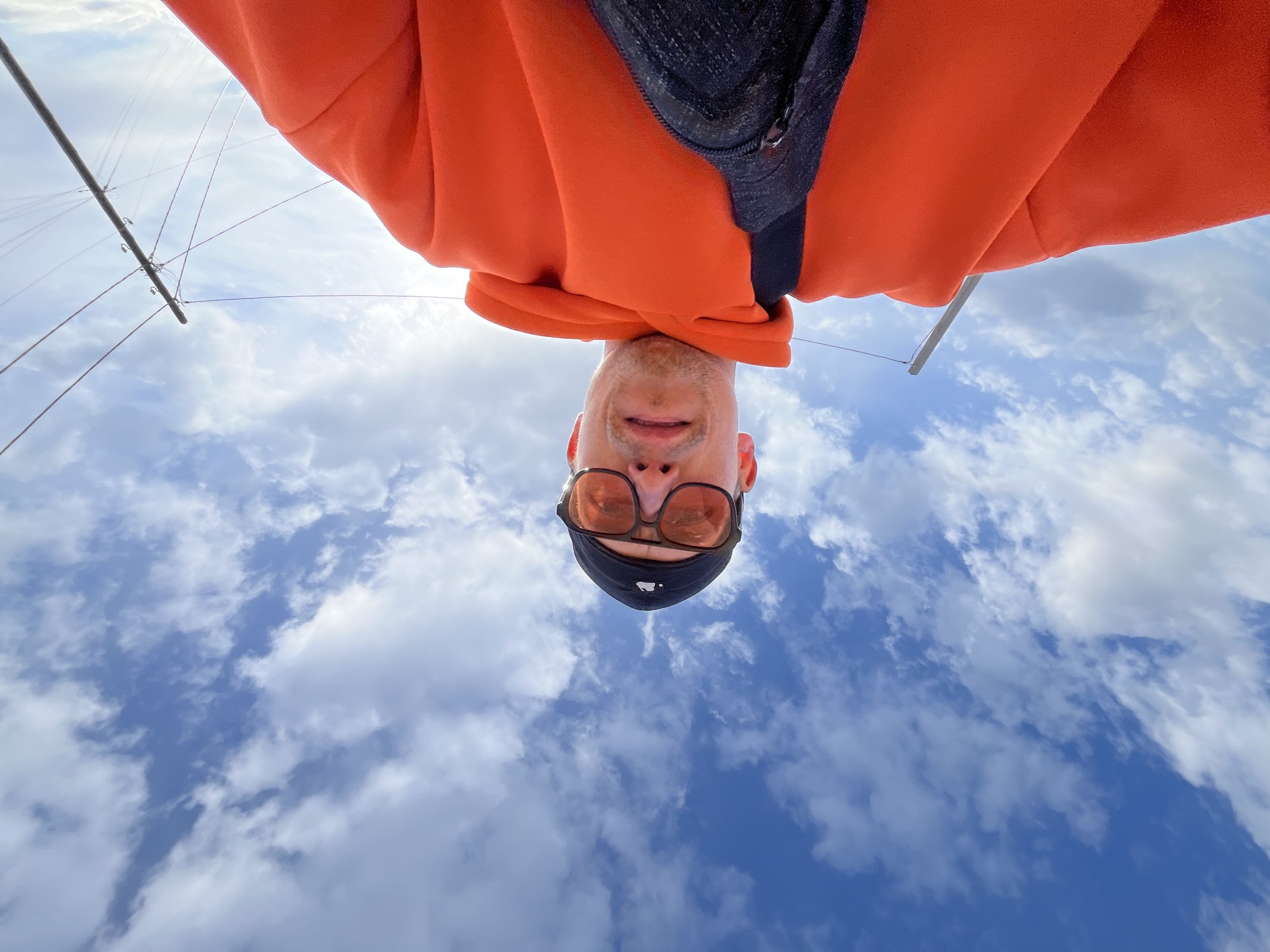 A low-angle selfie captures a person looking directly downwards (due to the image's inverted orientation) against a bright blue sky filled with scattered white clouds. The subject, likely male, is wearing an orange hoodie, a dark top underneath, sunglasses with reddish-brown lenses, and a black face covering or balaclava. The unique perspective makes the person appear upside down, as if looking up from below. Visible in the upper left corner of the frame are elements of a mast and rigging, including a metal pole with small hooks and thin lines, suggesting a marine or sailing environment. The orange fabric of the hoodie also partially frames the top right of the shot. A small, white, circular logo, resembling an Apple logo, is faintly visible on the black face covering below the nose. The scene depicts a bright, clear daytime with good visibility of the sky and clouds, indicating pleasant weather. The unusual angle gives the image a dynamic and creative feel, possibly taken on a boat or near water in Chaikovsky, Russia.