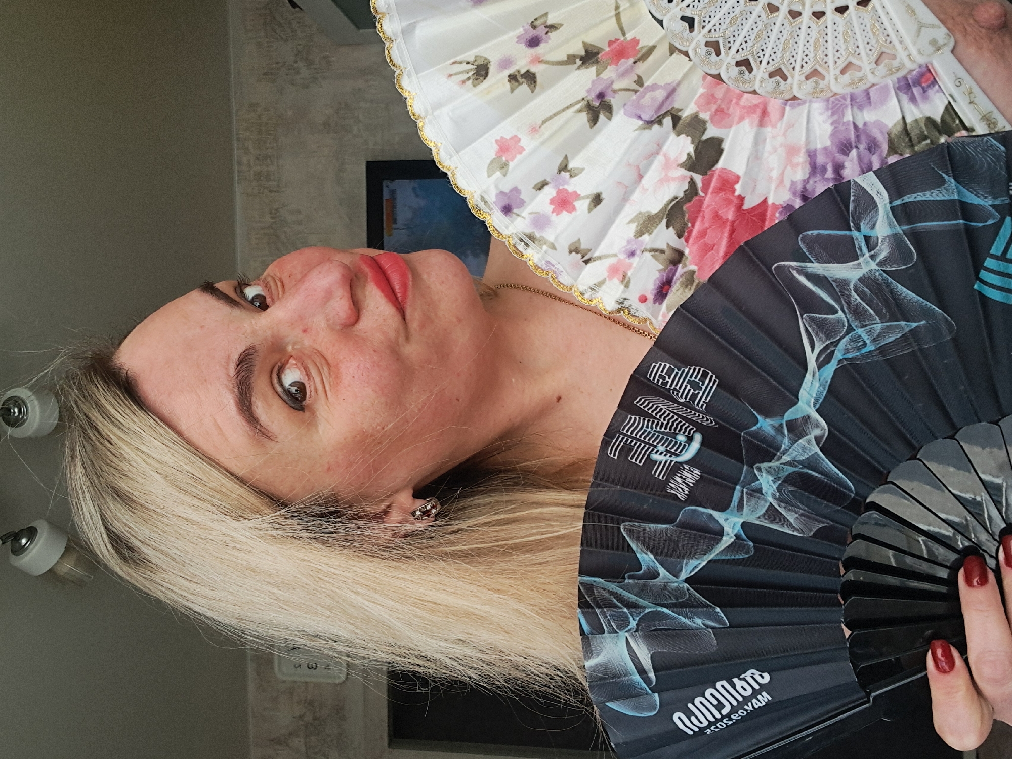 A fair-skinned woman with light blonde hair and red lipstick is holding two open fans in front of her face. One fan is black with a light blue pattern and writing in a non-English alphabet, while the other fan is white with floral patterns in pink, purple, and green, and trimmed with gold edging. The woman has an earring in her left ear. The background includes a wall with a textured pattern, light fixtures, and a framed object. The image may have been taken in Elektrostal, Russia.