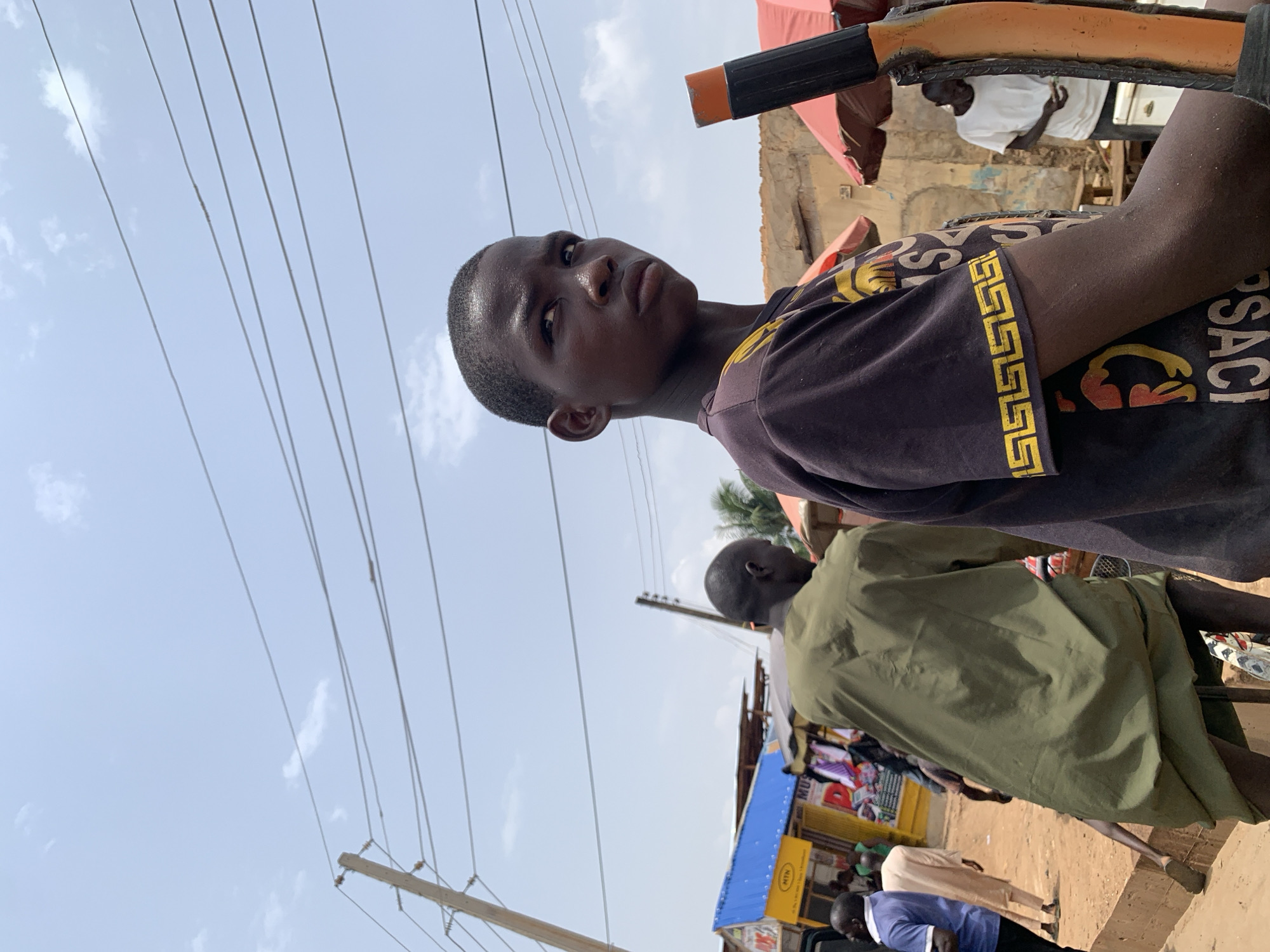 Akwanga, Nigeria. A young person with dark skin, short hair, and a black shirt with a gold pattern is visible in the foreground, looking upwards. They are holding a dark and orange object. Behind them and to the right is a person wearing a green shirt. In the background, partially obscured, are more people, a building with a blue roof, and an MTN sign. Overhead are several power lines against a light blue sky.