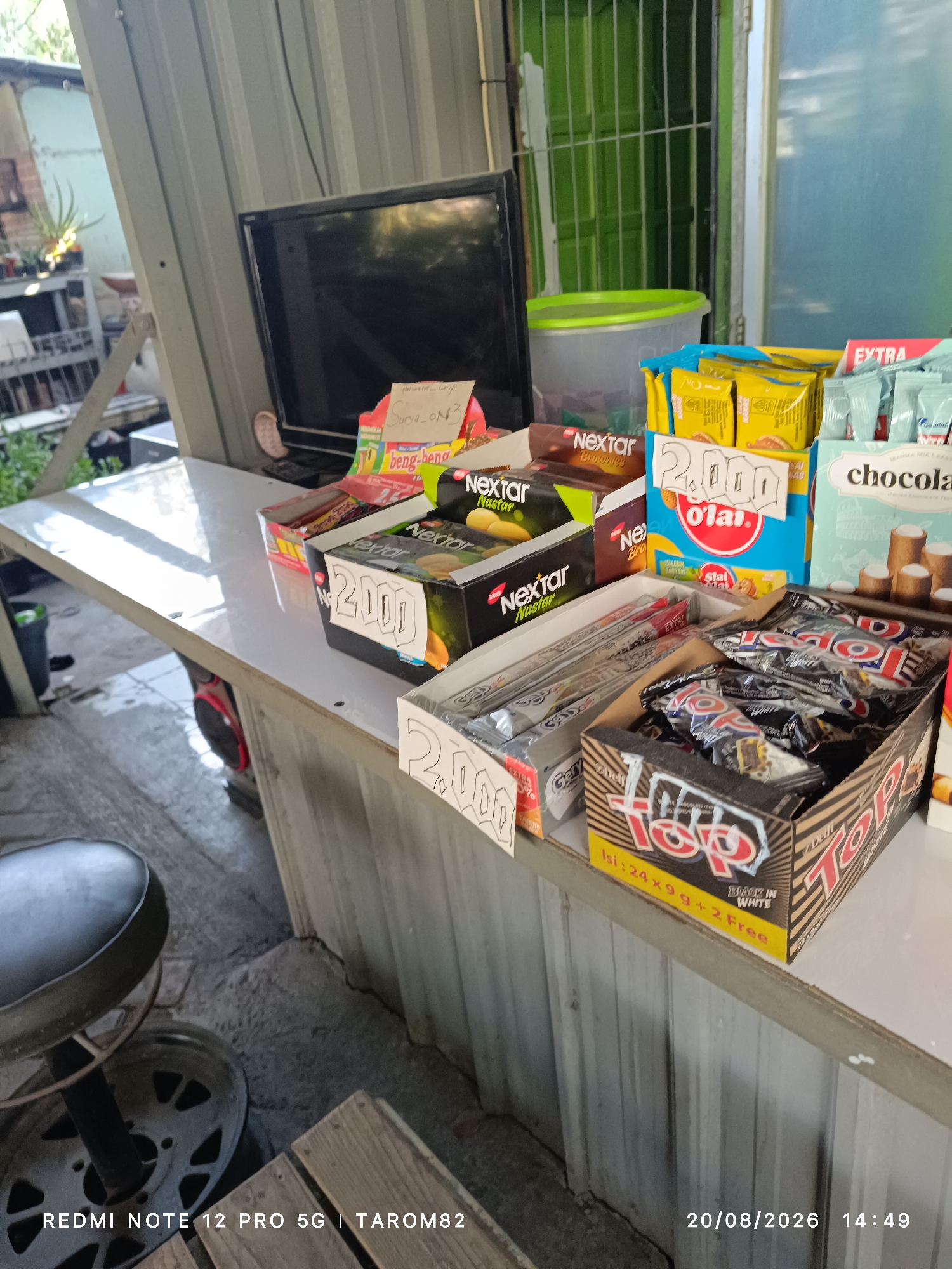 This is a medium shot photograph taken outdoors at a street-side vendor stall in Tebel, Indonesia. The stall is set up against a corrugated metal wall, with a white laminate countertop displaying various products for sale. In the foreground, on the left, a black padded stool with a metal base is visible.
The countertop is laden with an assortment of packaged snacks and sweets. Several boxes of "Nextar" brand cookies are prominent, some featuring chocolate and brownie flavors. Next to these, there are boxes of "O'lai" snacks and "Top" brand chocolate bars, with one box clearly labeled "Black in White". Some products have price tags, with "2.000" visible on multiple items. Behind the snacks, a black flat-screen television is mounted on the wall. To the right of the television, a transparent plastic container with a green lid is situated.
The background shows more of the corrugated metal wall and a glimpse of a green-grated door or window. The ground appears to be paved with concrete. The lighting suggests it is daytime, with sunlight casting reflections on the countertop. The overall impression is that of a small, informal retail setup selling confectionery and snacks.