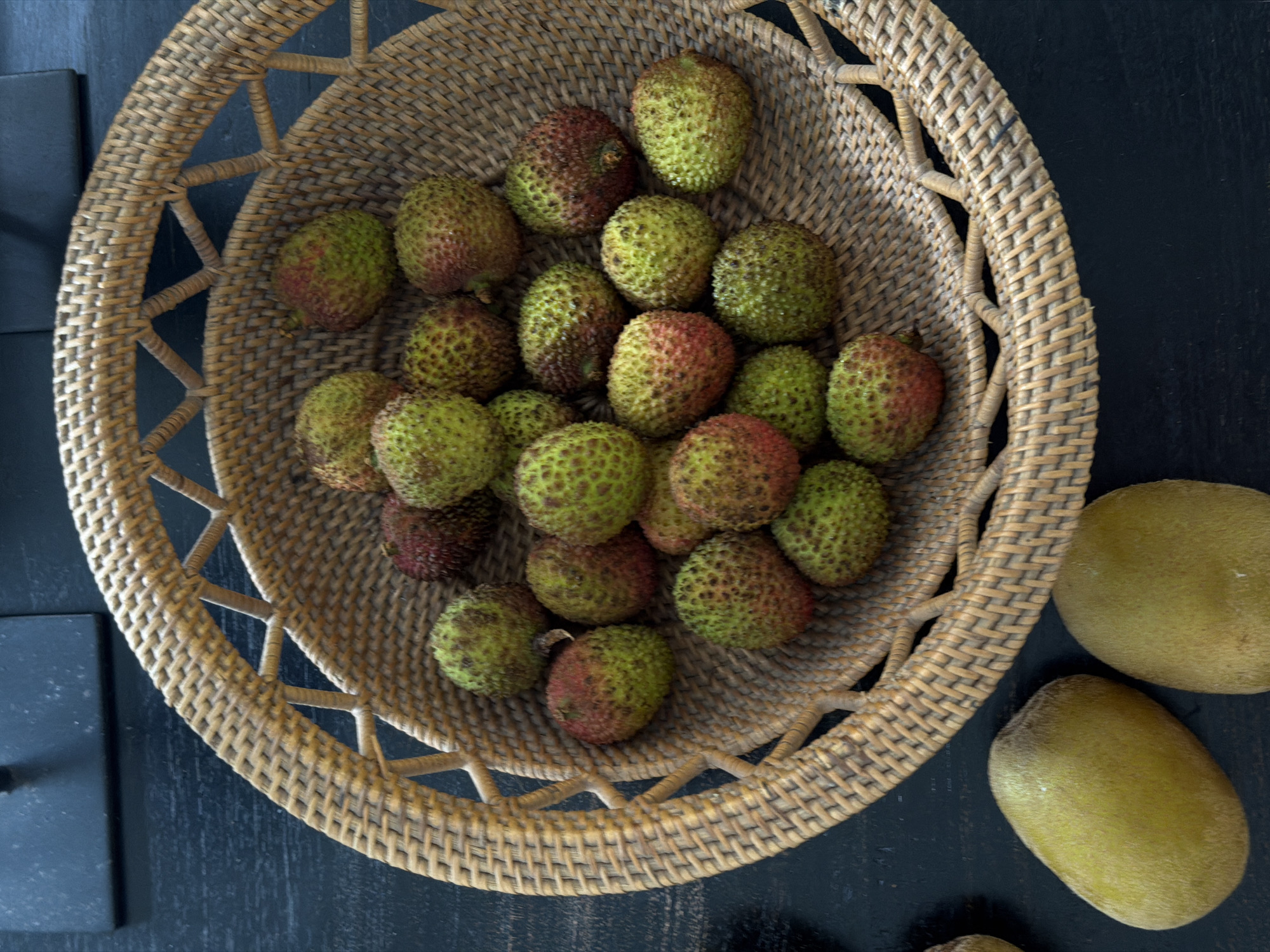 The central subject is a round, woven wicker basket brimming with numerous small, bumpy fruits. These fruits display a variegated coloration, transitioning from bright green to reddish-brown hues, characteristic of unripe or partially ripe lychees. Outside the basket, to the right, are two elongated, fuzzy, yellowish-green fruits, identified as golden kiwis, with a third partially visible at the bottom edge of the frame. All subjects rest on a dark, textured surface, likely a wooden table. A dark, rectangular object is partially visible on the left side of the frame.
The image captures a static arrangement of fresh fruits, presented as a still life. No specific activity or event is occurring.
The wicker basket is made of light brown material, showcasing a detailed, open weave pattern around its rim and a tighter weave for its base. The lychees' skins have a distinct, bumpy texture. The golden kiwis are smoother than their green counterparts, with a subtle fuzz. The underlying surface is a deep, dark hue, appearing as distressed or painted wood, providing a stark contrast to the lighter fruits and basket. The lighting is even and soft, coming from above, highlighting the textures of the fruits and the intricate weave of the basket. The photograph is taken from a direct overhead perspective. The location context is North Kuta, Indonesia. No visible text or overt emotions are present.