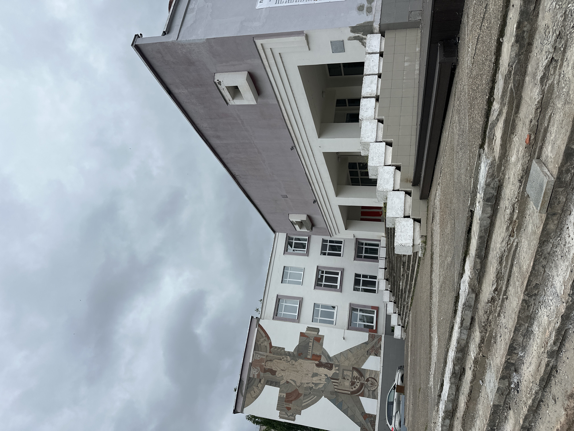 The image depicts the exterior of a building complex, likely a cultural or educational institution, located in Chaikovsky, Russia. The building on the left features a large, abstract mural with human figures and geometric shapes. Adjacent to it, a white building with multiple windows rises. To the right, a larger, gray building with a modern facade and a covered entrance area with a ramp is visible. The foreground shows a wide, outdoor staircase made of concrete, some sections of which appear worn and damaged. The sky is overcast with gray clouds. A silver car is parked in front of the muraled building, and several concrete planters are placed along the entrance. There are no people visible in the image.