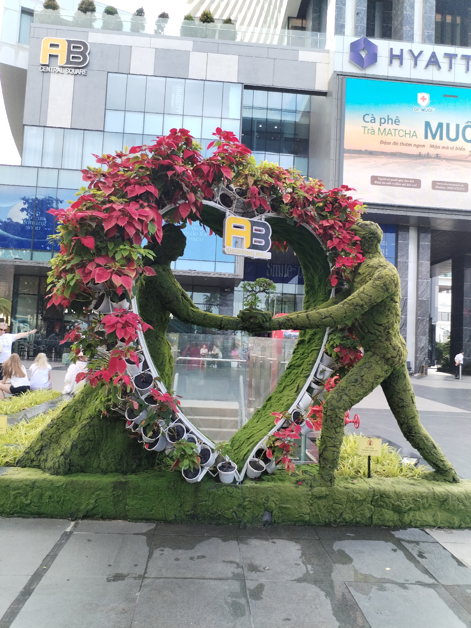 In Nha Trang Ward, Vietnam, an outdoor scene features a large, heart-shaped topiary sculpture constructed from green moss and foliage, adorned with red poinsettias. Two human-shaped topiary figures, one on the left appearing to be a woman and the other on the right a man, are intertwined within the heart shape, their moss-covered hands clasped. The figures hold a small bonsai tree between them. The sculpture is set on a raised, moss-covered base. In the background, a modern building with a prominent "HYATT" sign is visible, along with a large digital billboard displaying text in Vietnamese and images of coffee and matcha tea. The ground in front of the sculpture is paved with large, grey tiles, some of which are wet. Several people are seated on a tiered, yellow-green landscaped area to the left of the sculpture, while others are visible in the background, including one person standing near the building entrance on the right.