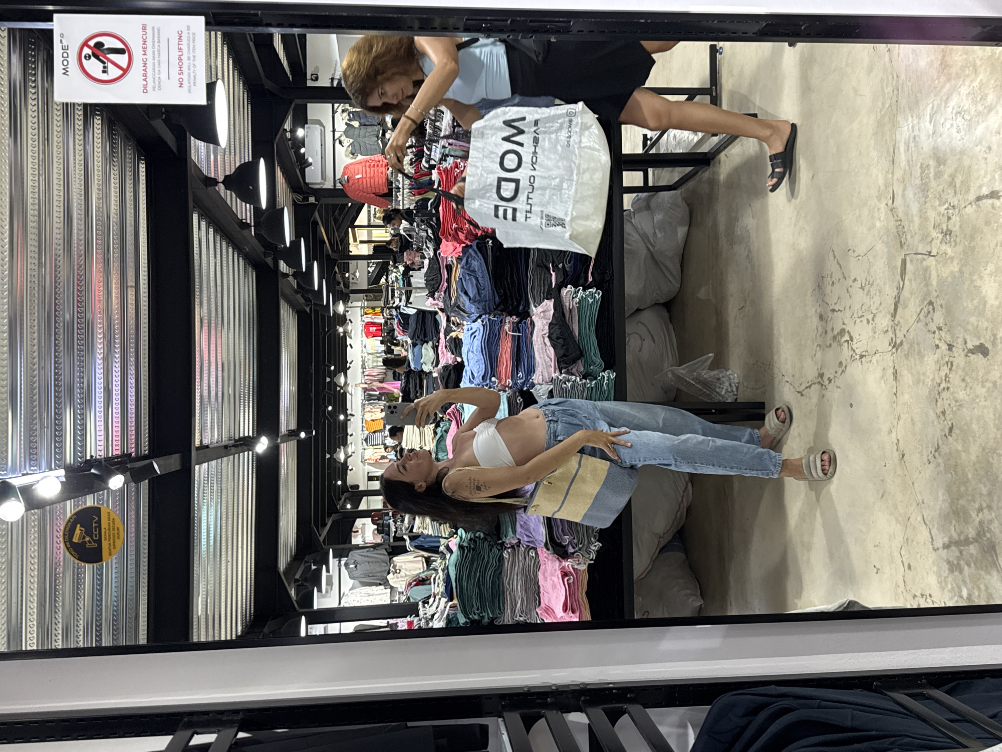 This is a vertical, full-shot photo taken inside a clothing store in North Kuta, Indonesia. The photo is taken from a low angle, looking up at a mirror that reflects the scene. Two women are visible. In the foreground, a woman with long dark hair and tattoos on her arm is taking a selfie in the mirror. She is wearing a white bandeau top and light-wash jeans, with a beige and blue straw bag slung over her shoulder. She is standing in front of racks of colorful t-shirts and apparel.
In the background, another woman with short brown hair is reaching for a white shopping bag with "MODE" printed on it in large, dark letters. She is wearing a light blue tank top and a black skirt. The store appears to be well-lit with overhead lights. Signs are visible on the wall behind the women. One sign with a red circle and diagonal line over a stick figure indicates "NO SHOPLIFTING" and includes text in Indonesian. Another circular yellow sign with a camera icon reads "CCTV." The overall atmosphere seems casual, suggesting a shopping trip. The time of day is not specified, but the indoor lighting suggests it could be any time.