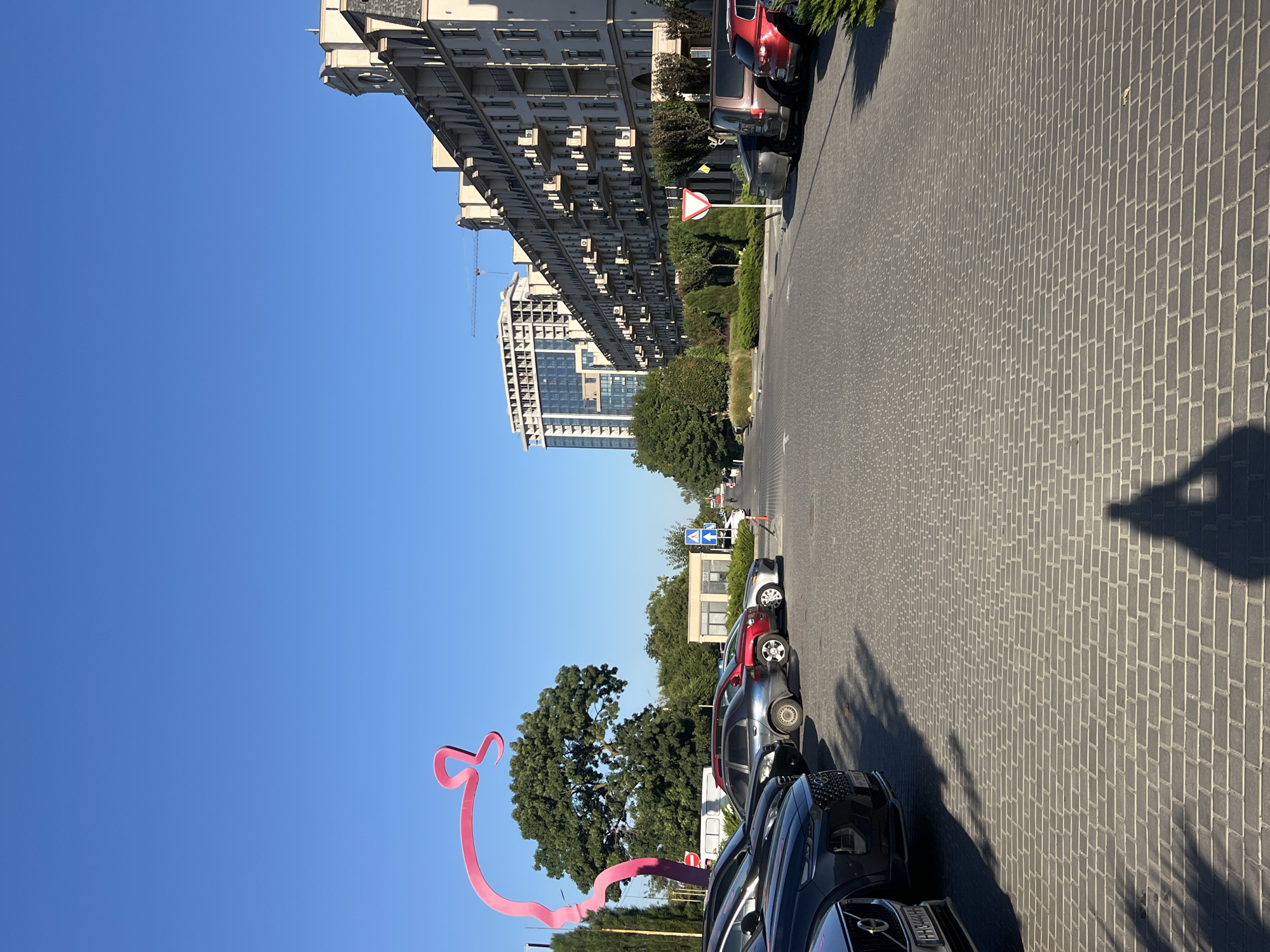 The image shows a street scene in Fontanka, Ukraine, on a clear day with a bright blue sky. In the foreground, a paved area with interlocking brickwork is visible, with several cars parked on the left side. A prominent pink sculpture resembling a flowing ribbon or abstract form is situated to the left of the frame, against a backdrop of green trees and a tall residential building under construction in the distance. To the right, a long, multi-story building with many balconies lines the street. Traffic signs are visible, including a yield sign. The shadow of a person or people is cast on the paved ground in the foreground.