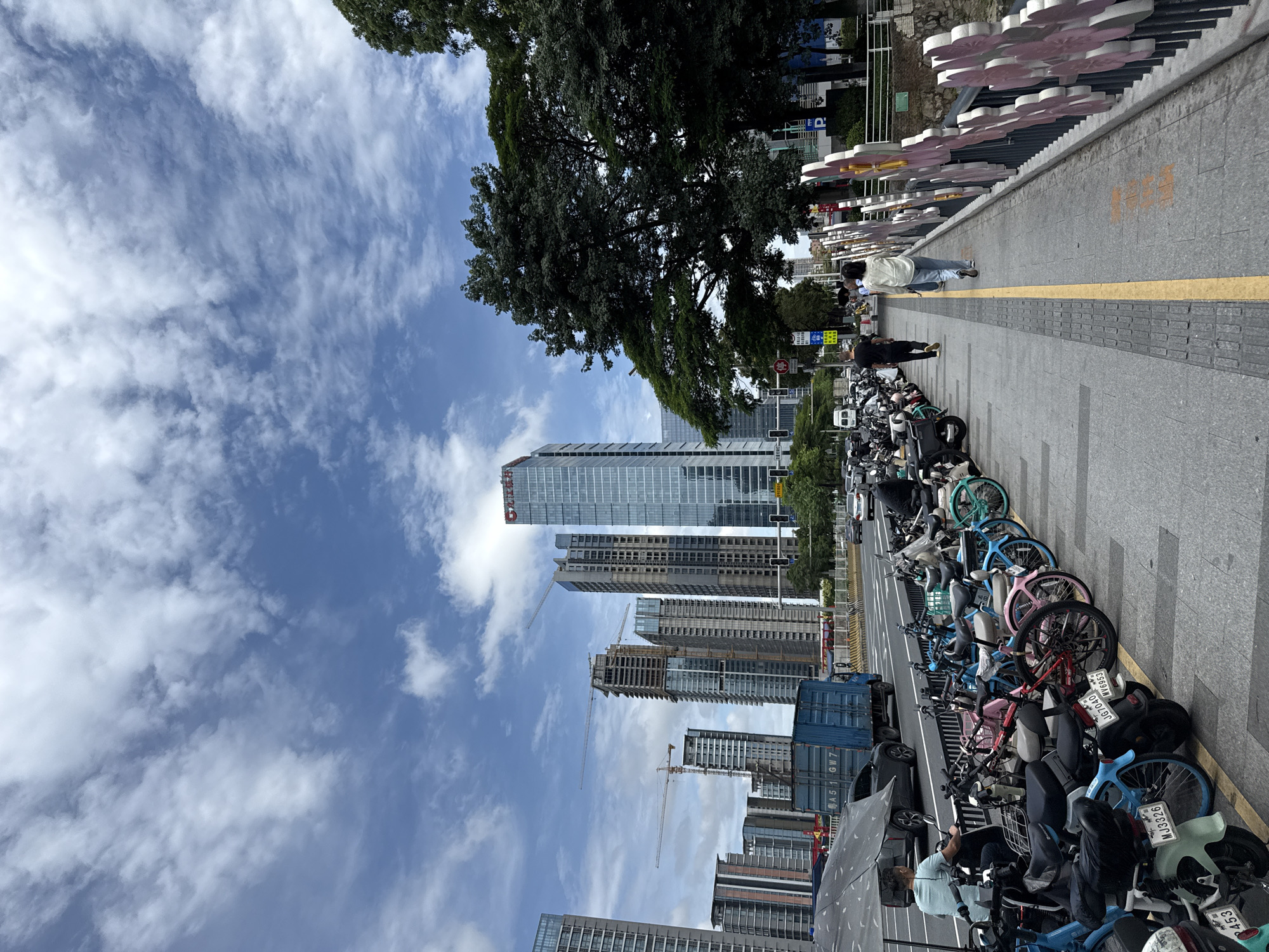 The image shows a street scene in Tianhe District, China, with a row of parked electric scooters and bicycles on the left. A man is seated on one of the scooters, wearing a light blue t-shirt and dark pants, with an umbrella over him. Further down the street, a blue container truck is visible. In the background, several tall buildings are under construction, interspersed with completed skyscrapers. The sky is blue with scattered clouds. On the right side of the image, a walkway is lined with a decorative metal fence featuring colorful floral designs. Two individuals are walking on the walkway, one in a white shirt and light-colored pants, and another in a black shirt and dark pants. A large tree with dense foliage is also present on the right. Traffic, including cars and a white van, can be seen on the road.