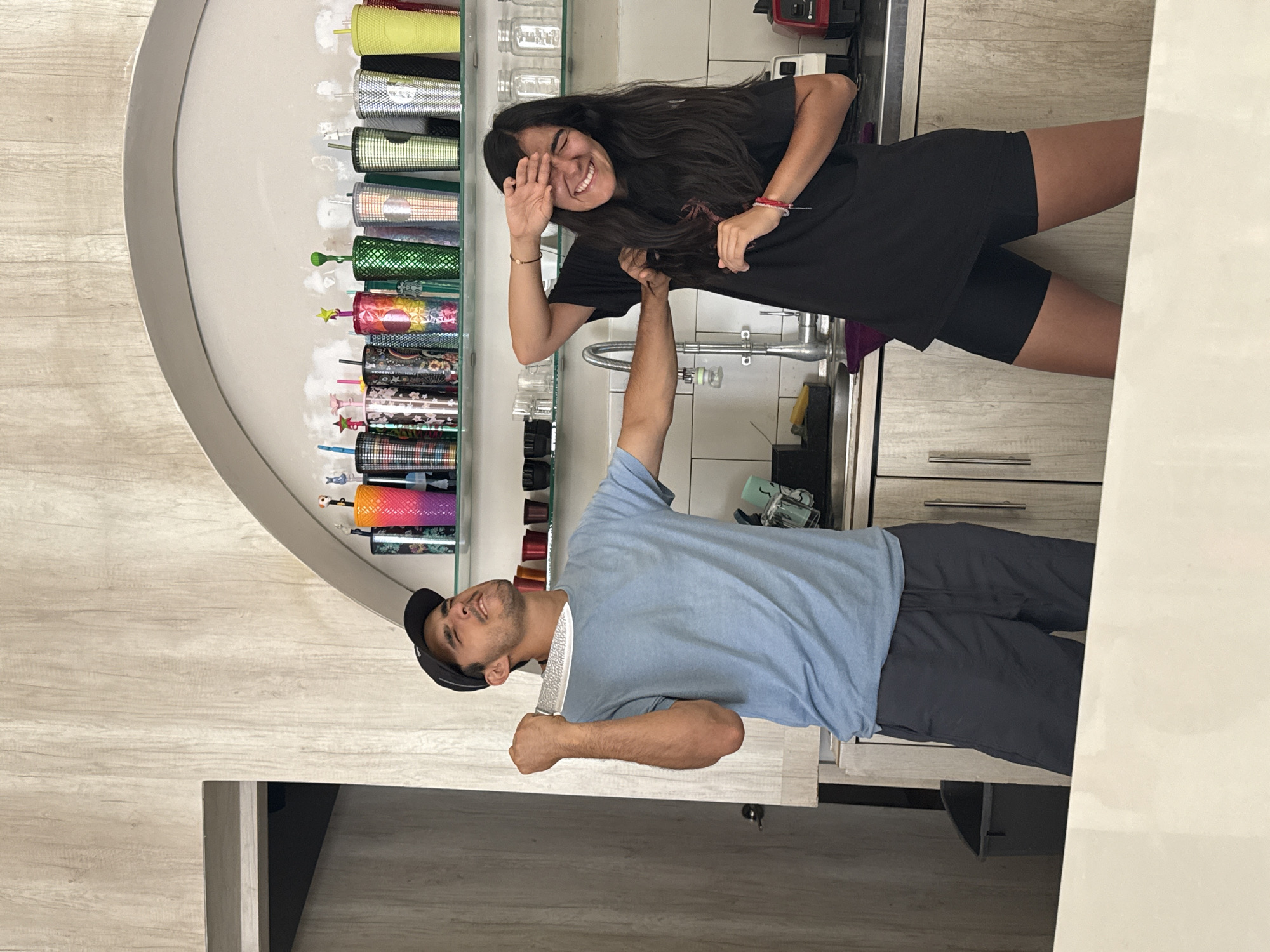 Two people are posing in a kitchen. A woman with long dark hair is wearing a black t-shirt and black shorts, with bracelets on her wrist. She is smiling and holding her hand up to her face. A man is wearing a light blue t-shirt, dark pants, and a baseball cap. He is holding the woman's hand in his raised right hand. A kitchen sink is in the background between them. There is a shelf of colorful tumblers behind the woman. The location is Puebla, Mexico.