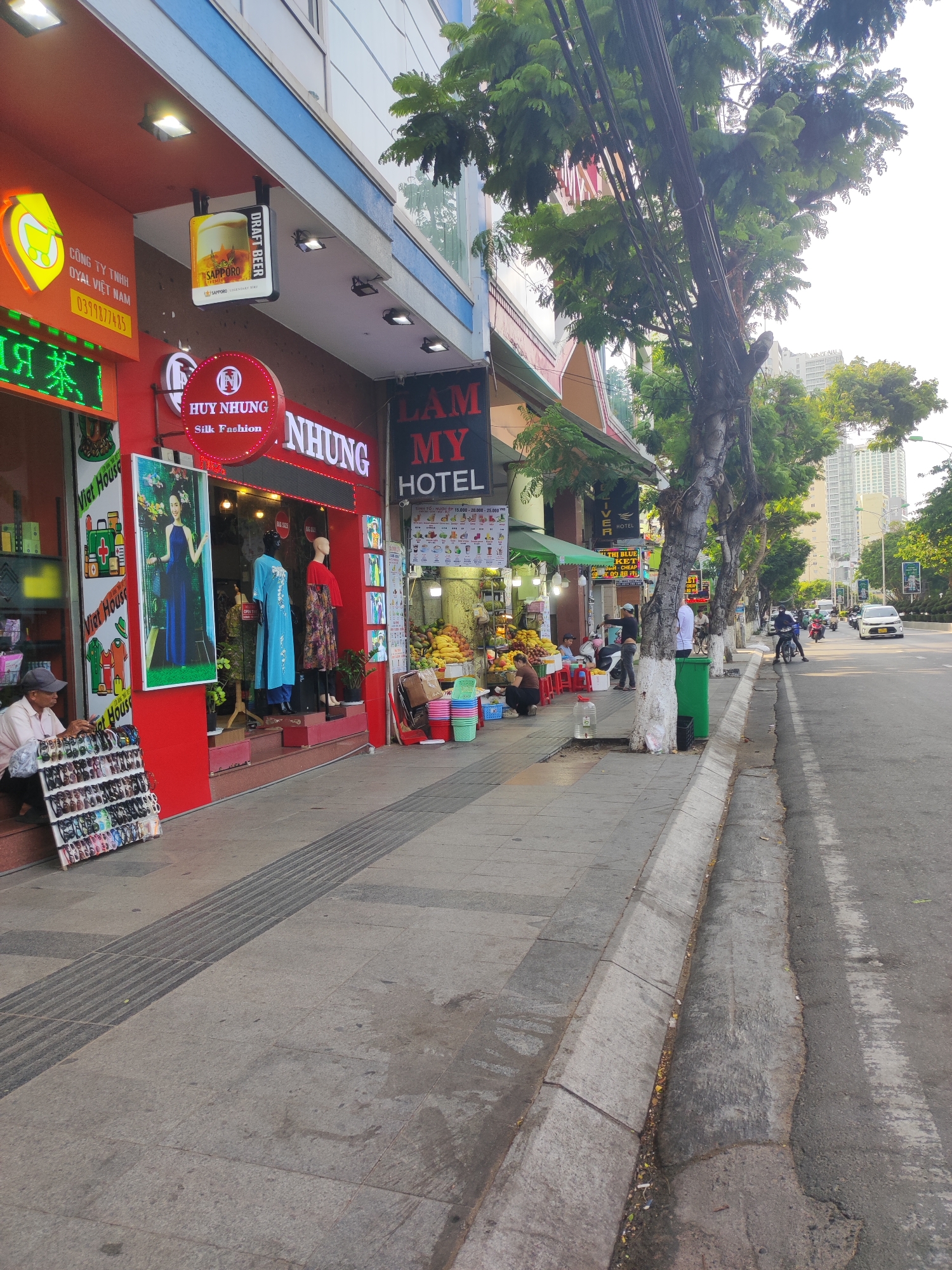 The image depicts a street scene in Nha Trang Ward, Vietnam, on a sunny day. The foreground shows a sidewalk with a textured, raised tactile paving strip. To the left, a row of shops is visible, with awnings and illuminated signs. One shop, "Huy Nhung Silk Fashion," features mannequins displaying clothing, and a poster of a woman in a blue dress. Another shop has a neon sign with Chinese characters and a display of various items, including what appear to be shoes and sandals arranged on a rack in front of the shop. A person is seated on the sidewalk next to this display.
Further down the street, more shops line the sidewalk, including one labeled "Lam My Hotel." A fruit stand with a display of various fruits like bananas and mangoes is set up on the sidewalk. Several people are milling about the fruit stand and sitting on red plastic stools.
The street itself is paved and has a curb. Traffic is visible, including motorcycles and a white car. Trees with dense green foliage are planted along the sidewalk, their branches extending over the street. In the background, tall buildings are visible, suggesting an urban environment. The sky is bright and appears slightly hazy.