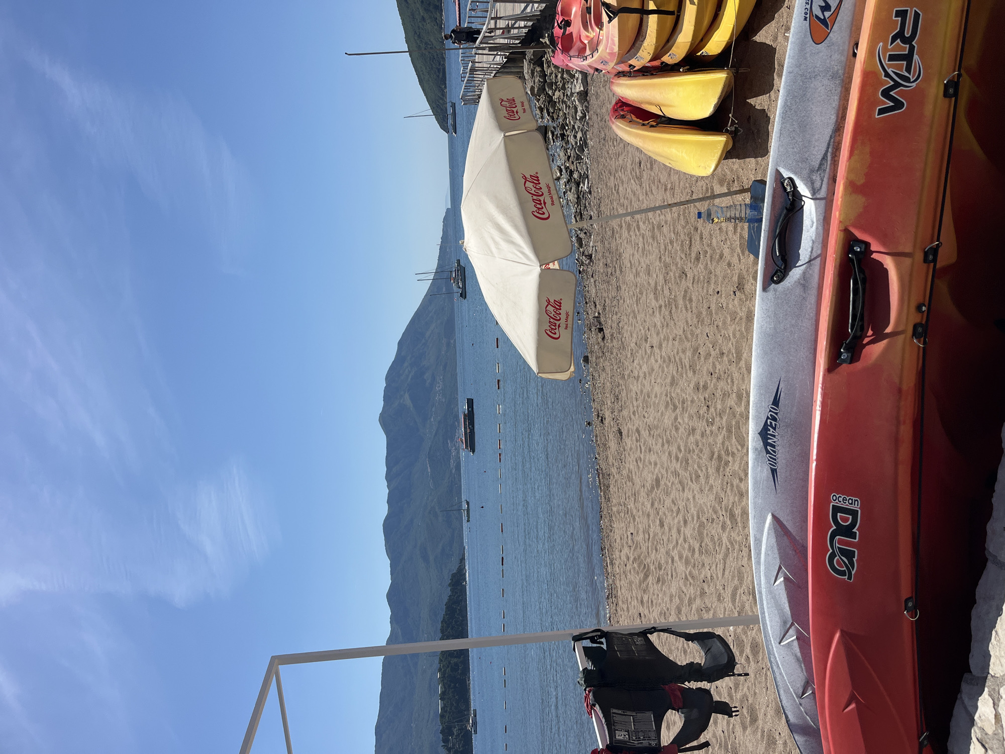 Here is a detailed description of the image:
The image shows a coastal scene at a beach, likely taken in Budva, Montenegro, given the presence of mountains and clear water. The foreground features a sandy beach with stacked kayaks of various colors including yellow, orange, and gray. A Coca-Cola branded beach umbrella stands near the kayaks, providing shade.
The middle ground includes the sea with boats anchored or passing by. In the background, a large mountainous landscape dominates the view, rising steeply from the coastline. The sky is clear and blue, with some light clouds visible.
Additionally, a structure, possibly a lifeguard stand frame, is visible in the lower left of the image, with life jackets hanging on it.